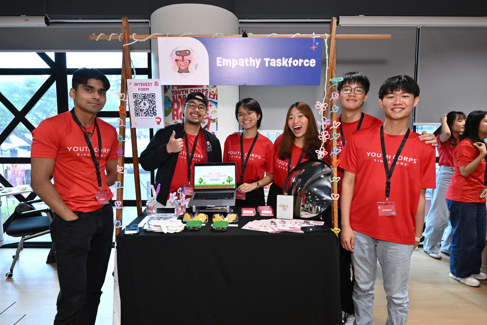 Youth Corps Singapore members at a booth, "Empathy Taskforce," with a sign and interest form.