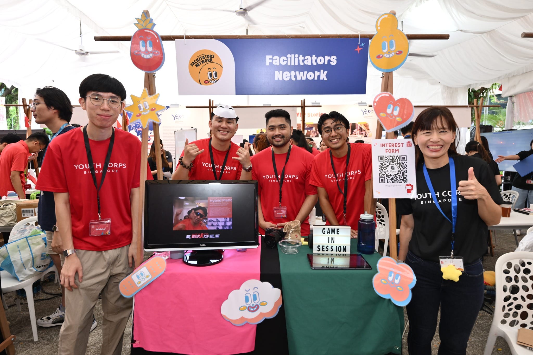Youth Corps Singapore booth: People in red shirts stand behind a table with monitors, signs, and cartoon cutouts.