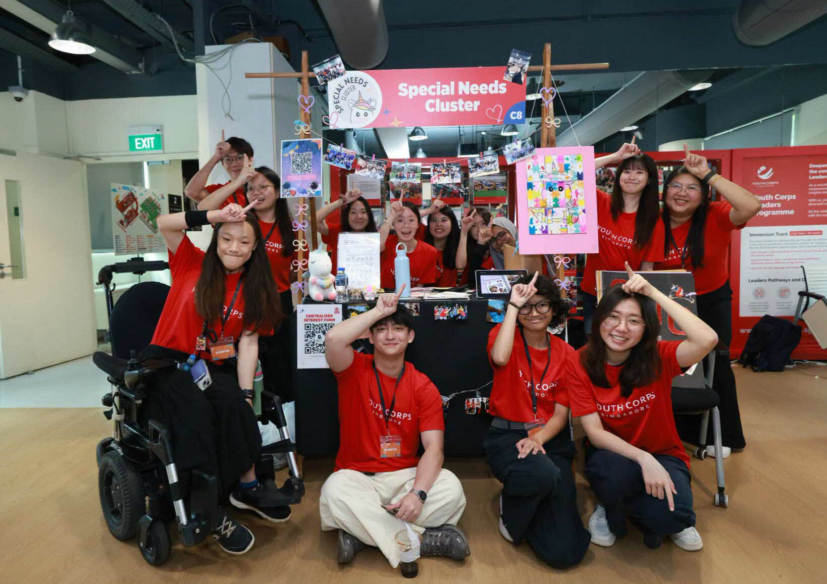 Group at "Special Needs Cluster" booth, Youth Corps Singapore, making unicorn signs. One person in wheelchair.
