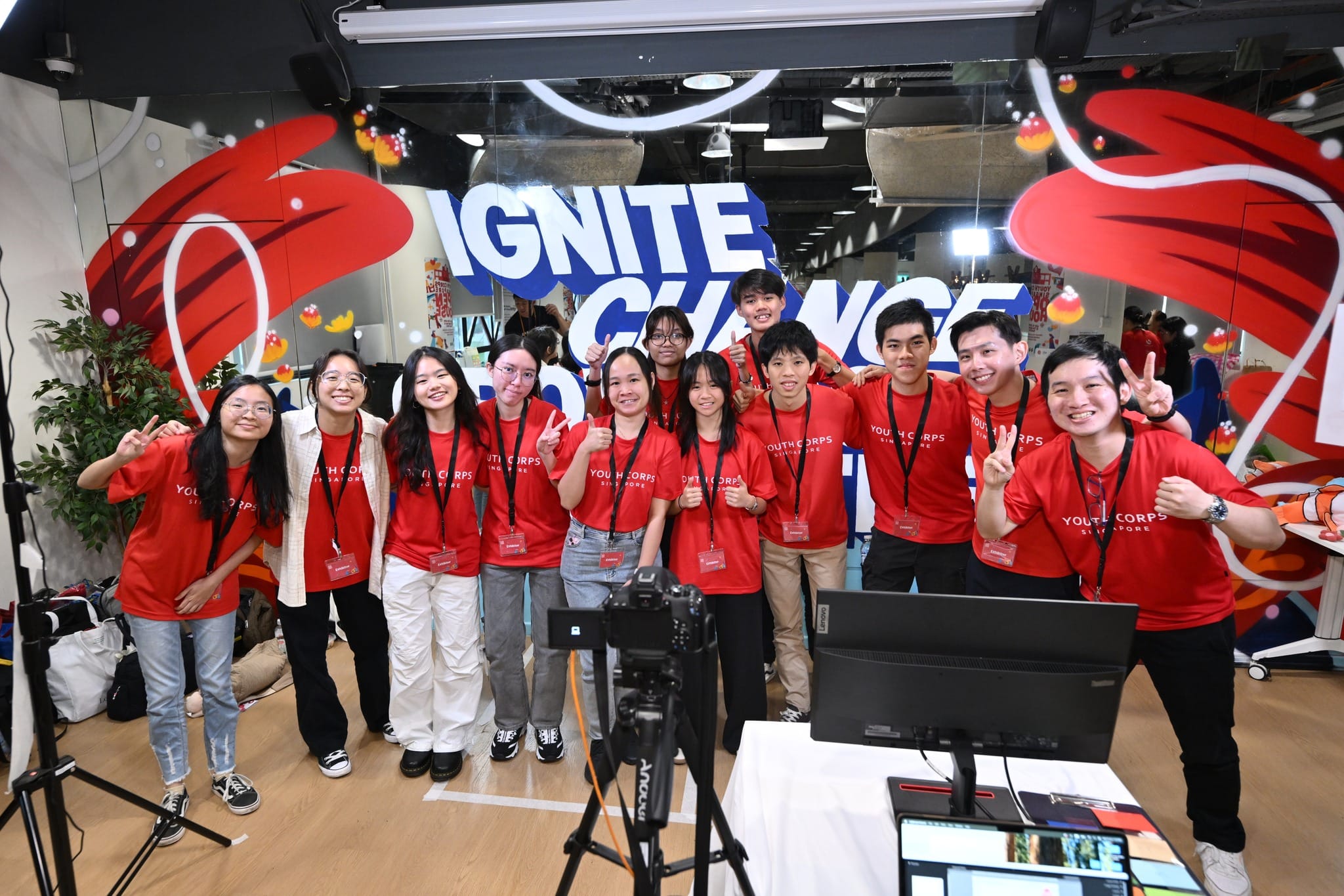 Group in red "Youth Corps Singapore" shirts with "Ignite Change" backdrop, posing for camera.