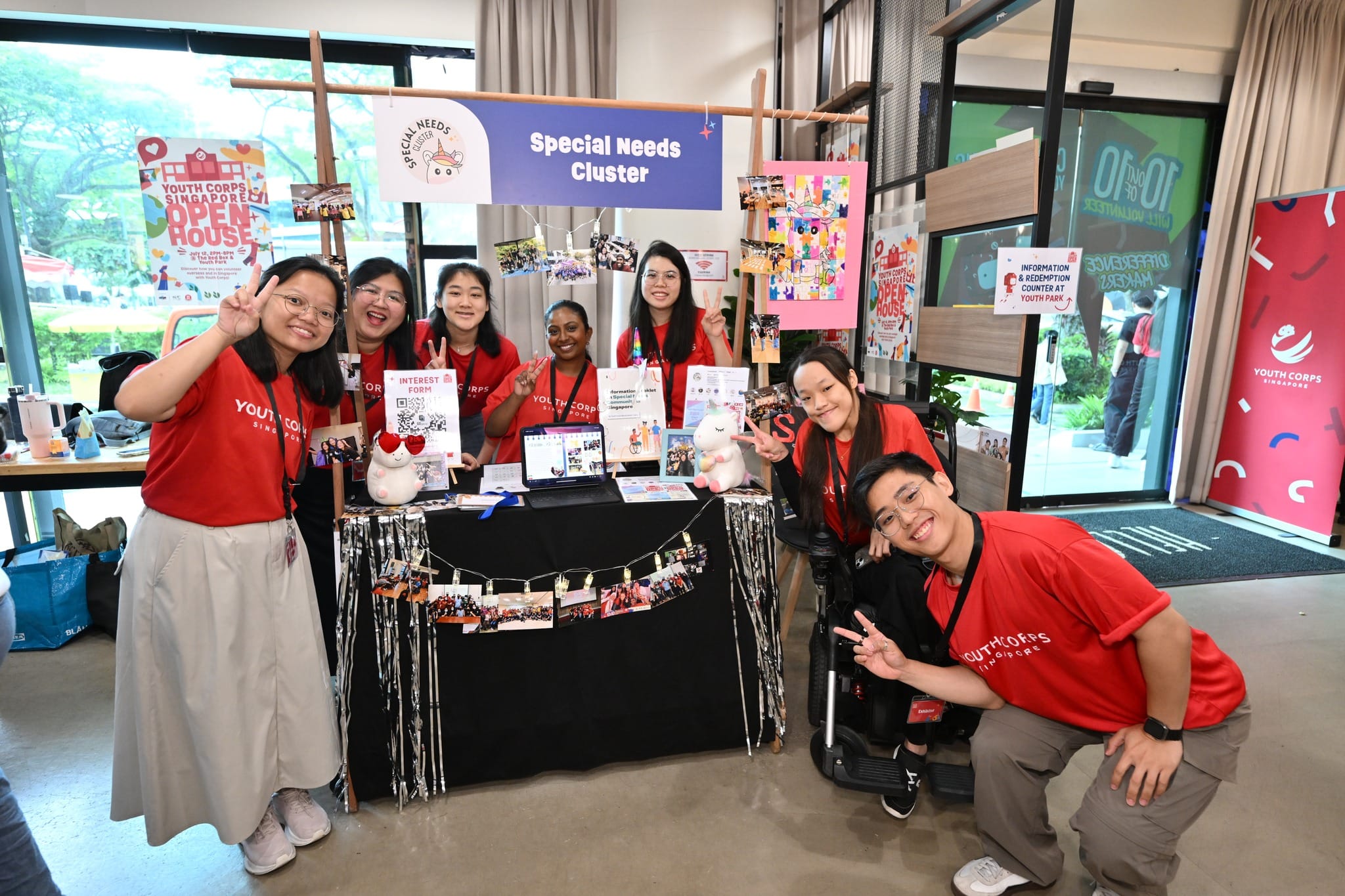 Group at "Special Needs Cluster" booth, Youth Corps Singapore, making peace signs. One person in wheelchair.