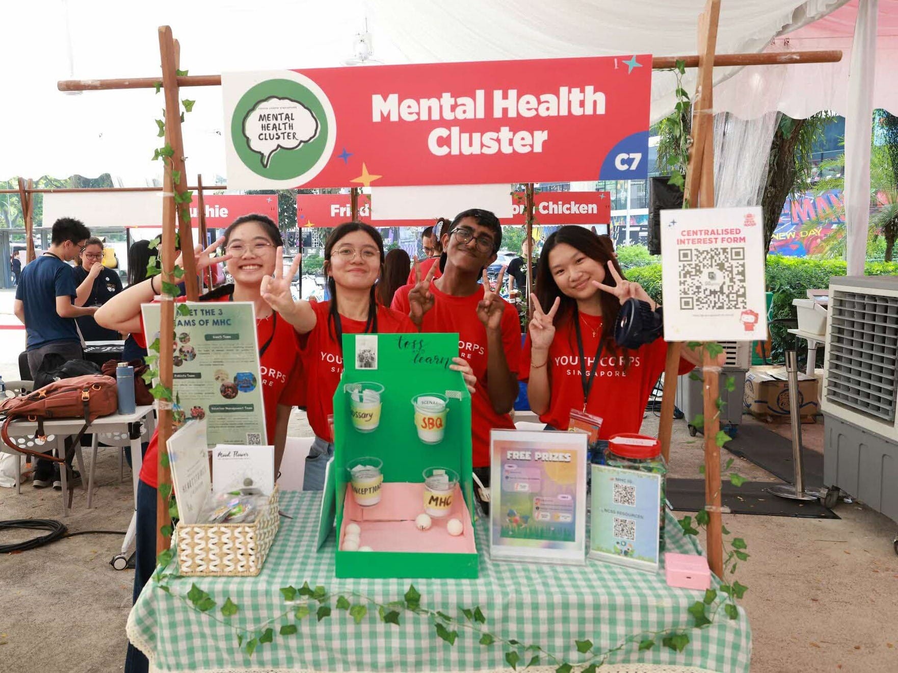 Group of young people in Youth Corps Singapore shirts posing in front of a "Mental Health Cluster" display.