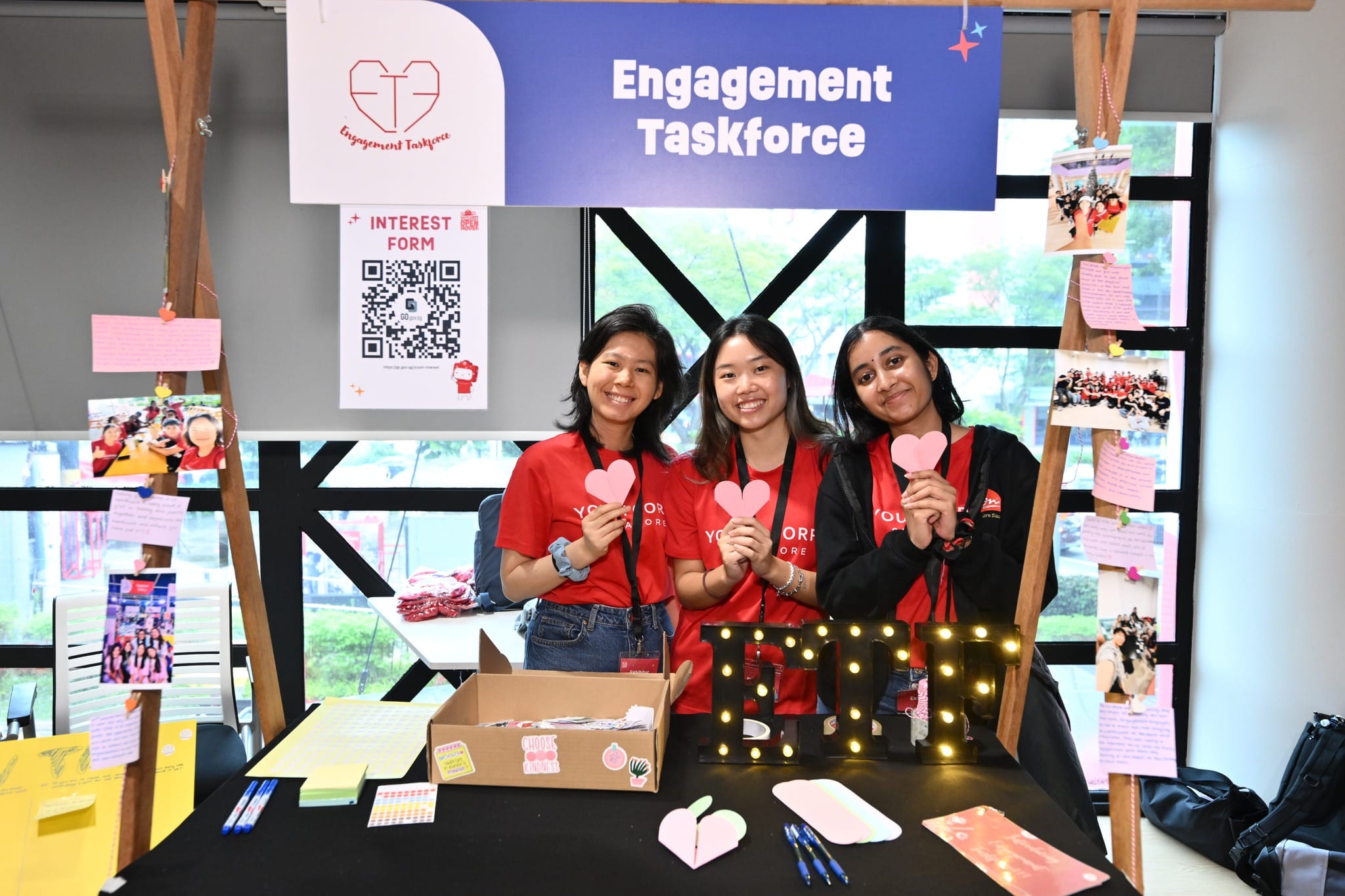 Three women at "Engagement Taskforce" booth, holding pink hearts, with a QR code interest form.