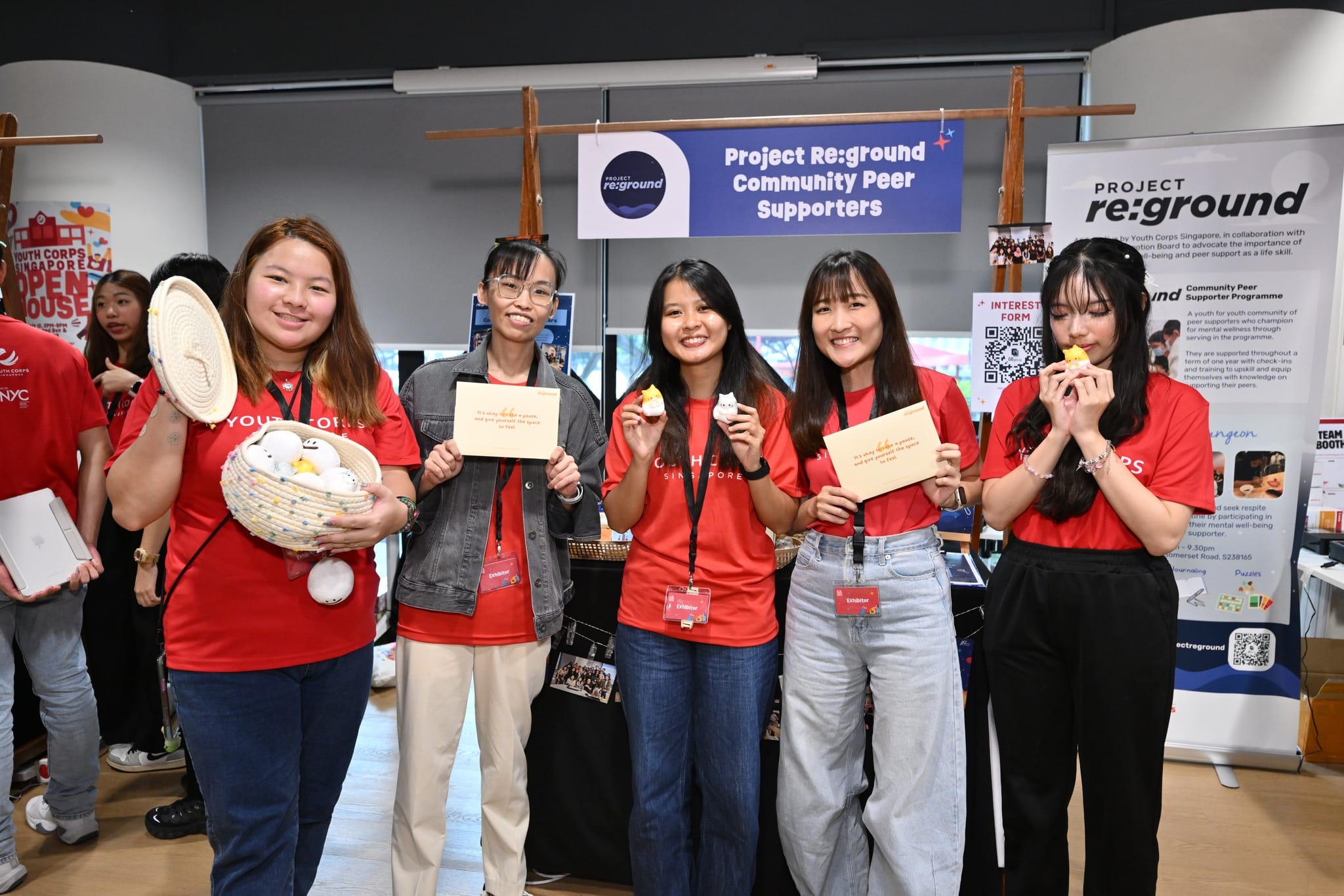 Five people at a Project Re:ground booth holding items, including a basket of toys and printed signs.