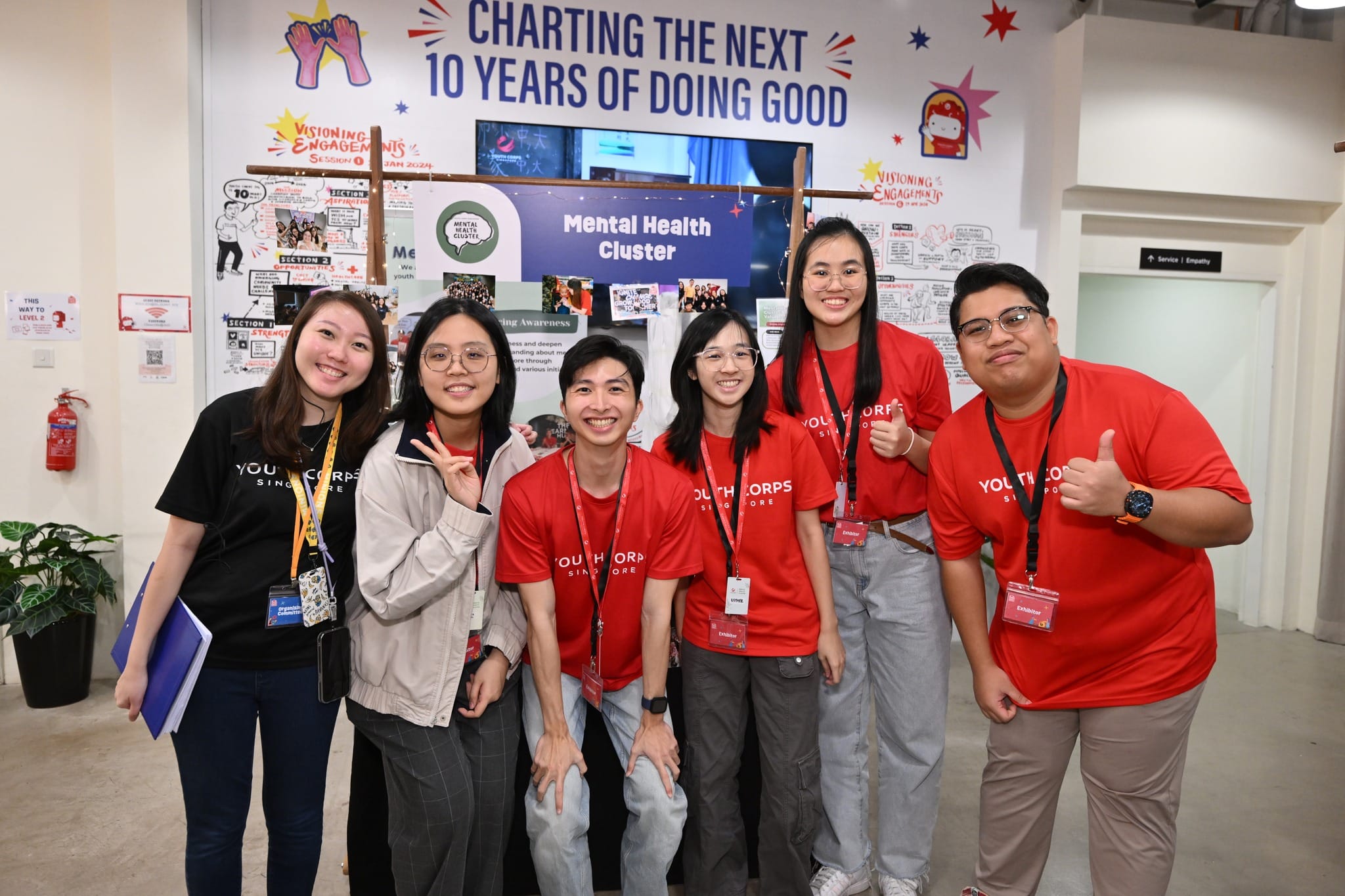 Group of young people in Youth Corps Singapore shirts posing in front of a "Mental Health Cluster" display.