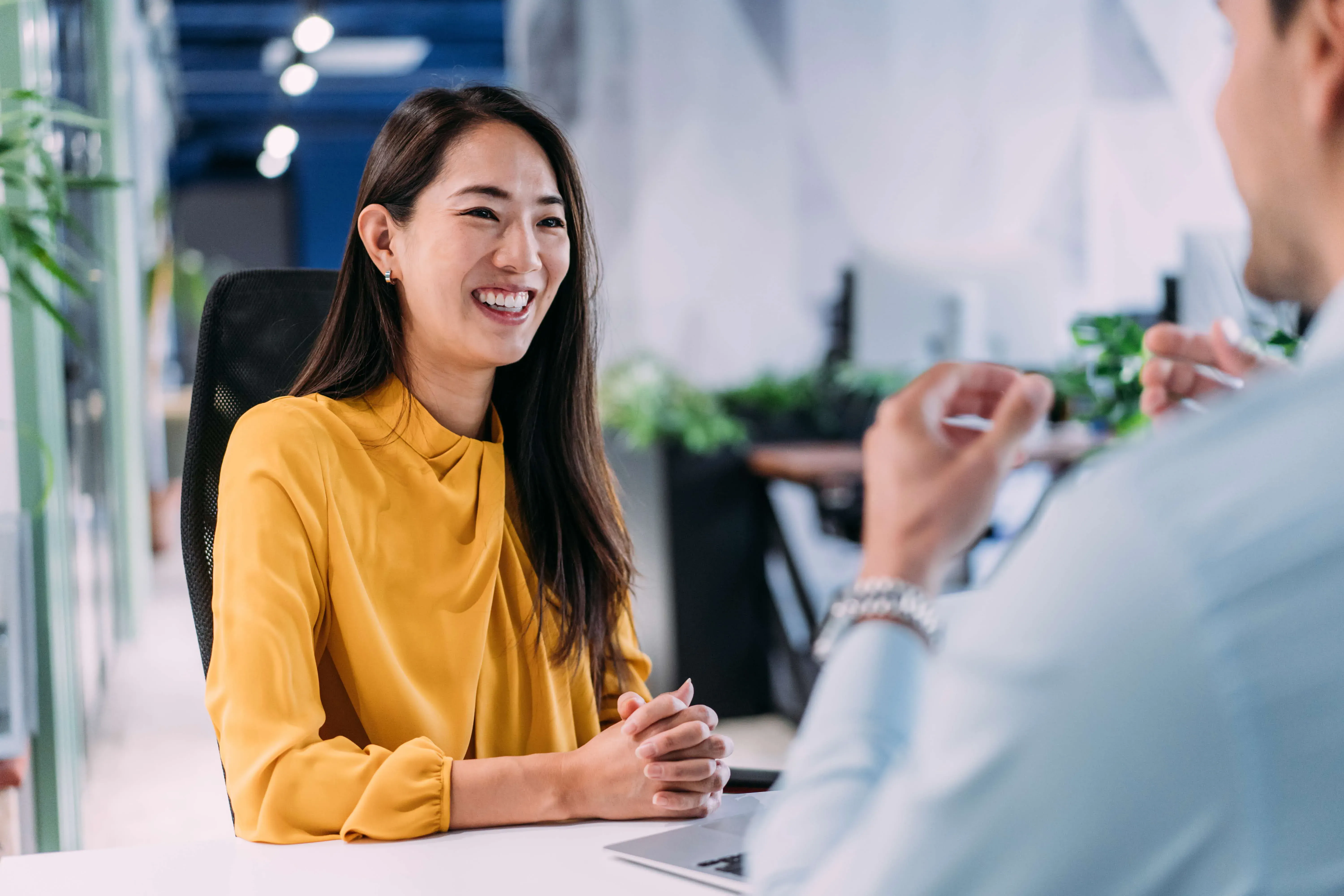 Smiling woman in yellow top sitting at desk in office meeting with person across table.