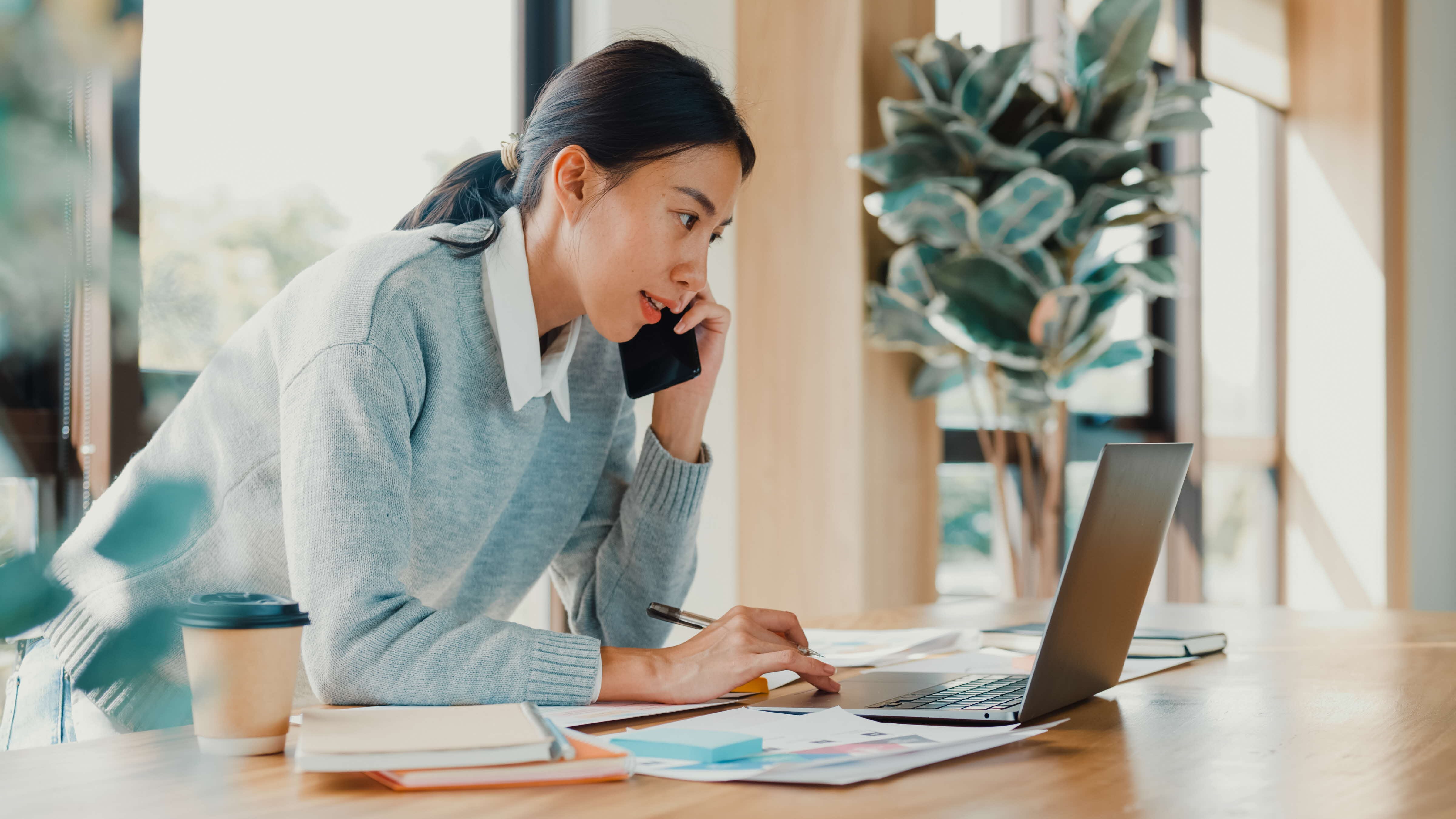 Woman in gray sweater talking on phone while working at laptop with papers and coffee.
