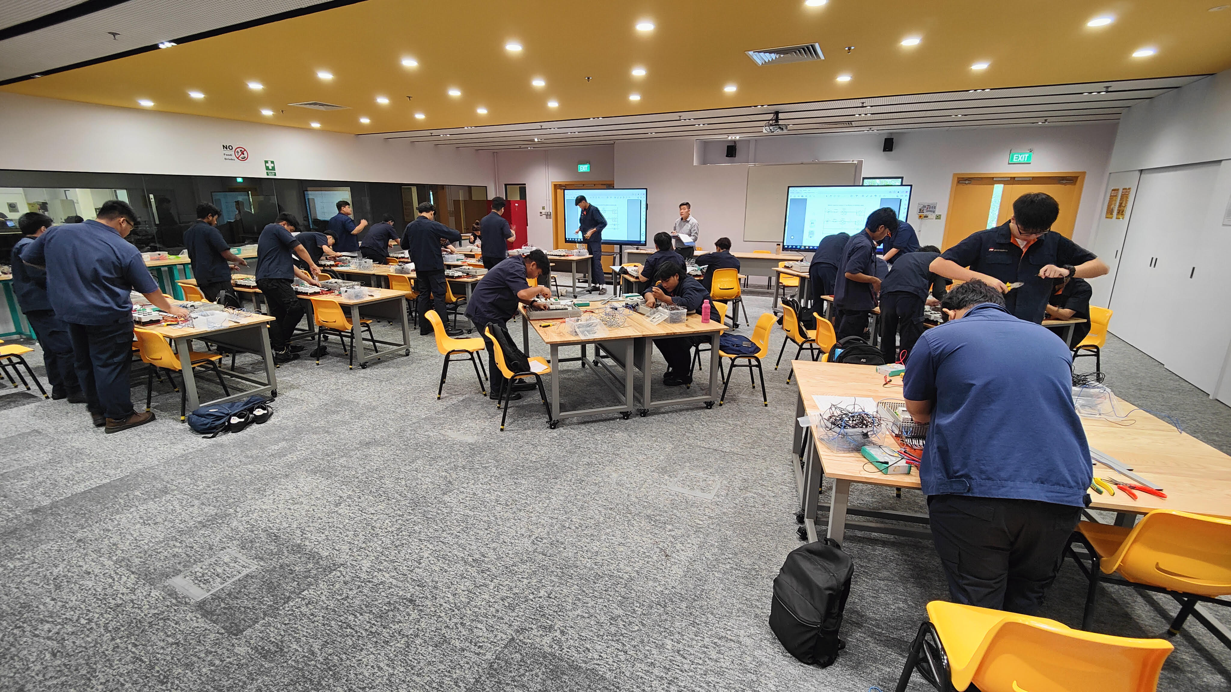 Classroom with students in blue shirts at tables working on electronics projects. Monitors display diagrams.