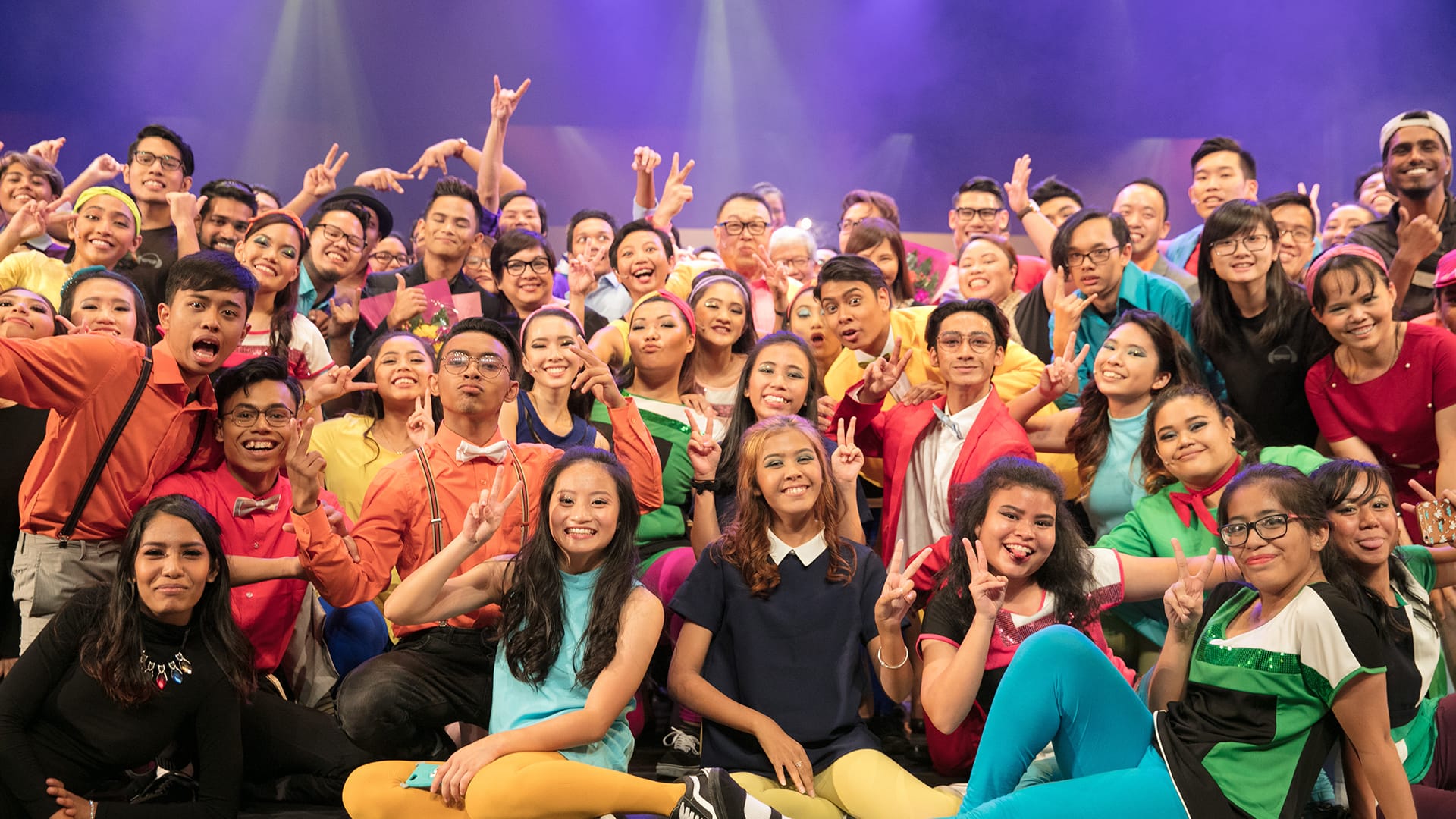 Large group of diverse people posed on stage, many making peace signs.