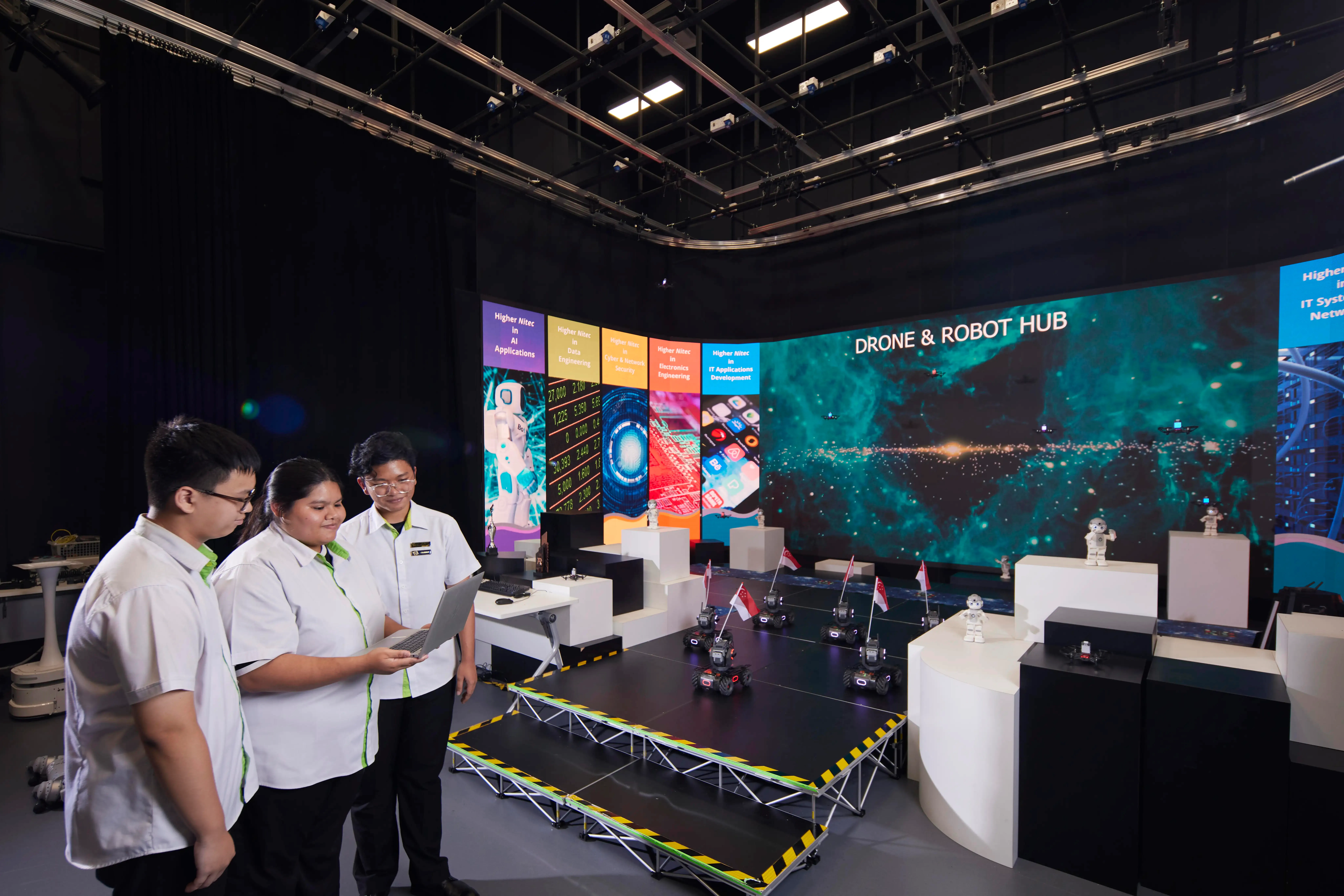 Students with laptop, drones with flags, robots displayed. /'Drone & Robot Hub/' on large screen.