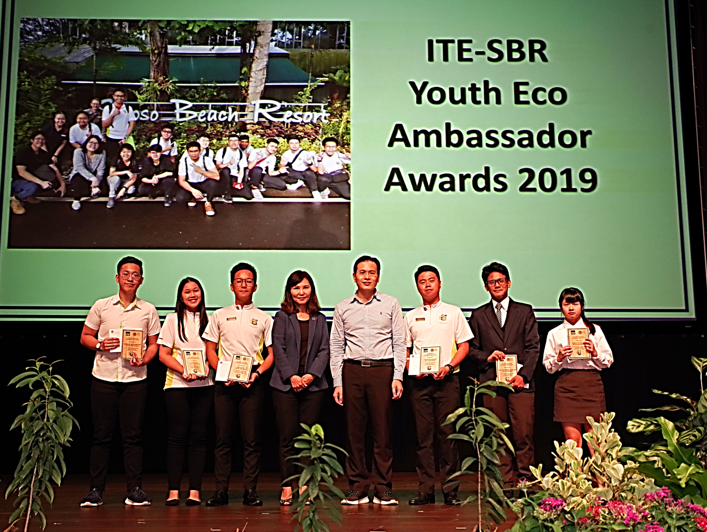 Eco Ambassador Awards 2019 ceremony: Eight people holding certificates on a stage with floral decorations.