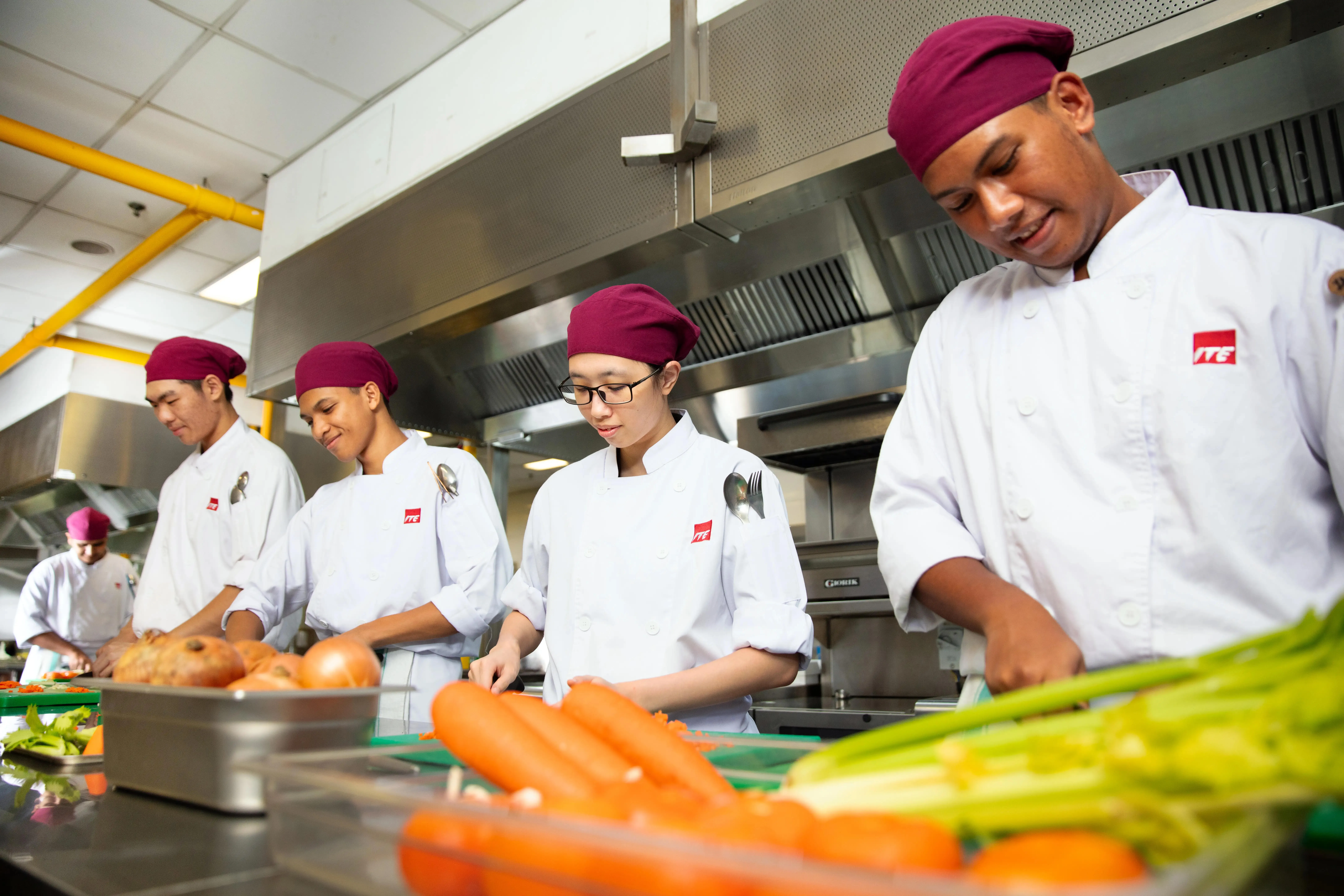 Chefs in white coats and maroon hats prep vegetables in a kitchen. ITE logo visible.