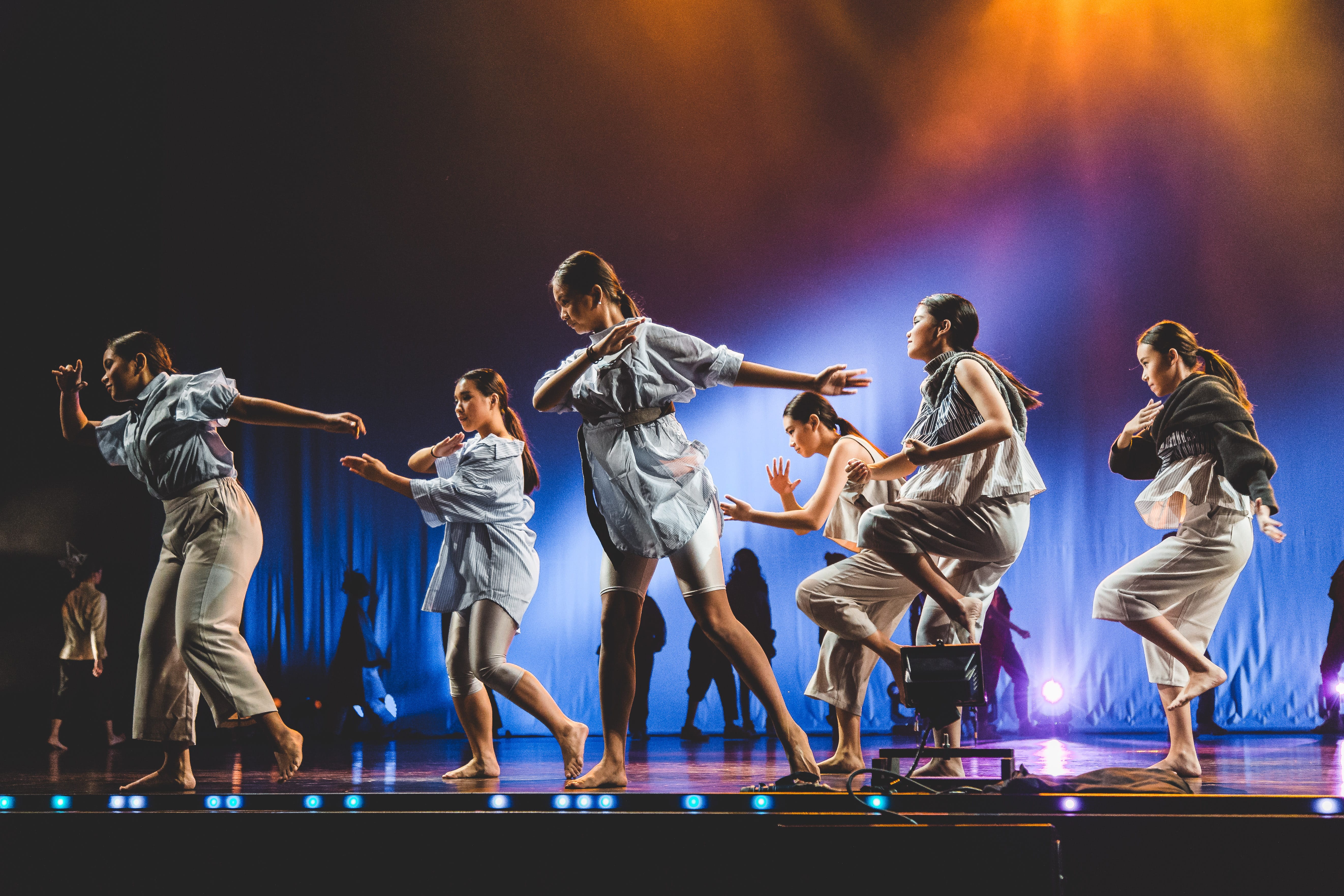 Dancers in loose, light-colored clothing perform barefoot on a stage with colored lighting.