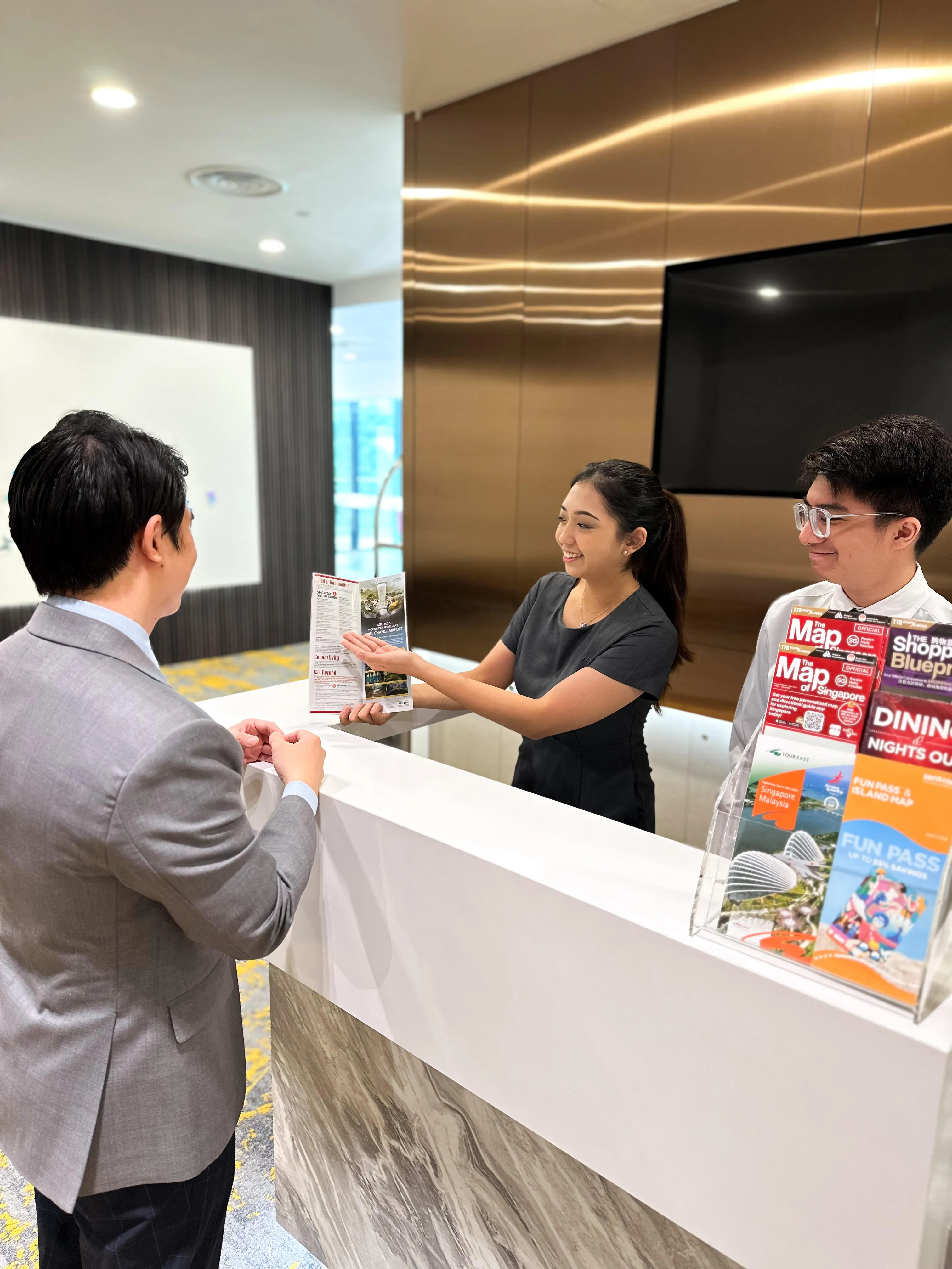 Reception area with two workers helping a guest; maps and tourist information are displayed.
