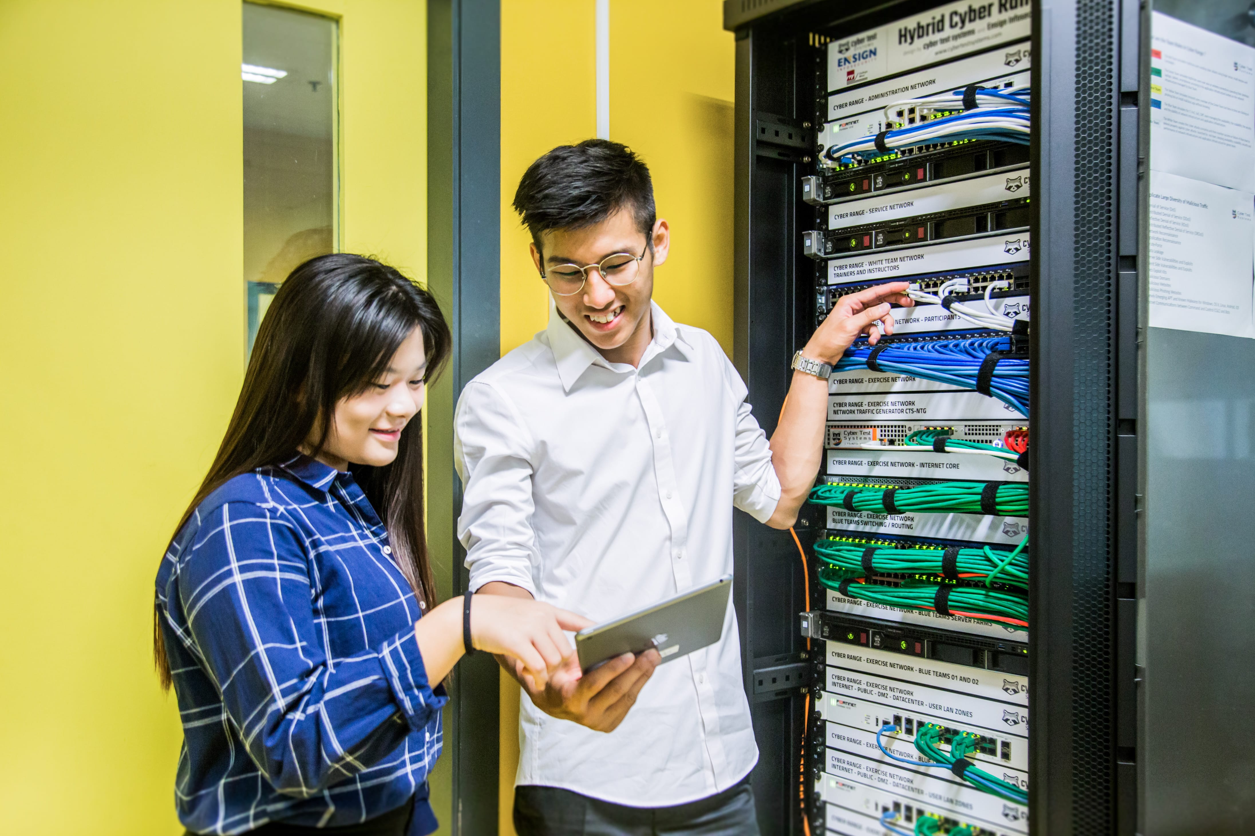 Man and woman examining tablet beside data centre server racks with labelled cables