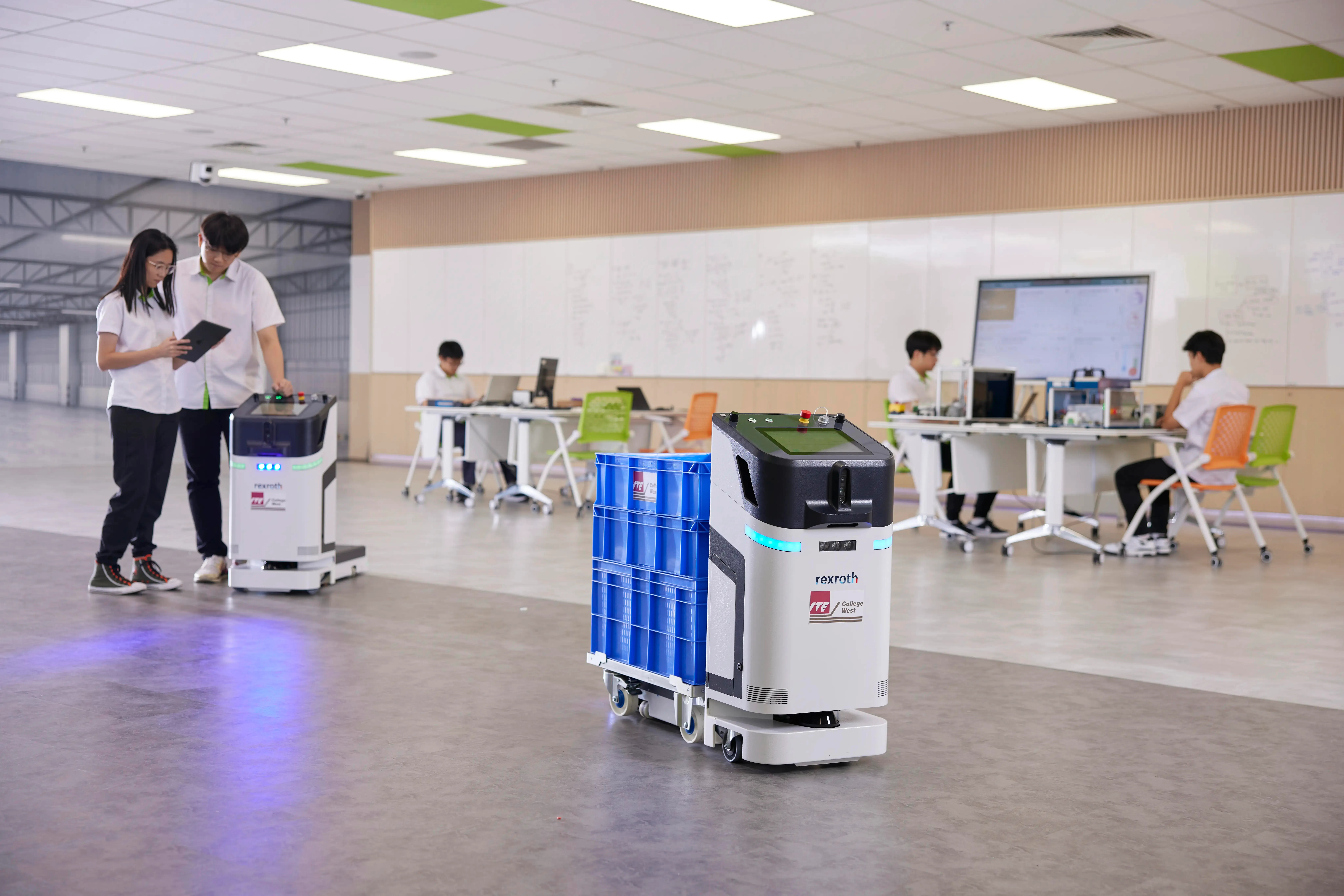 Rexroth robots with blue bins move through a room with students at desks.