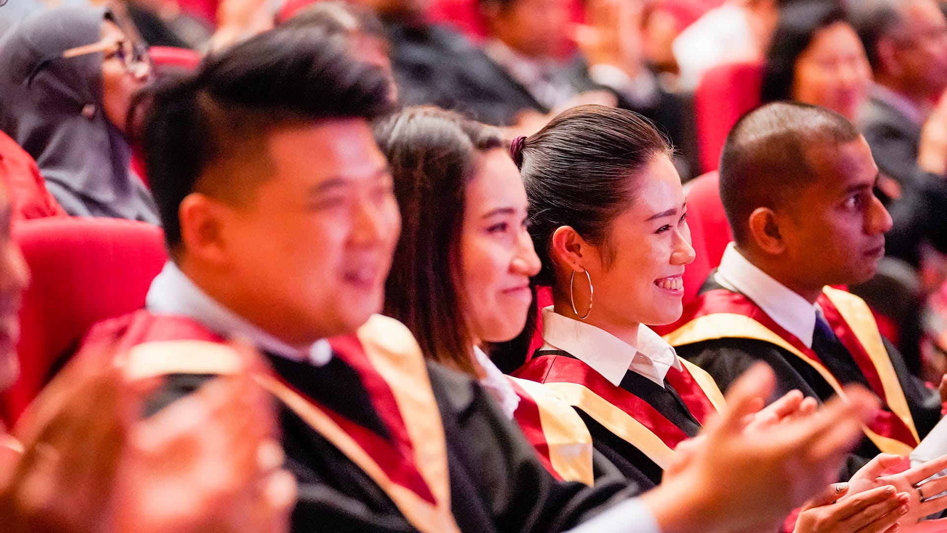 Graduates in black gowns with red and gold stoles clapping at a commencement ceremony.
