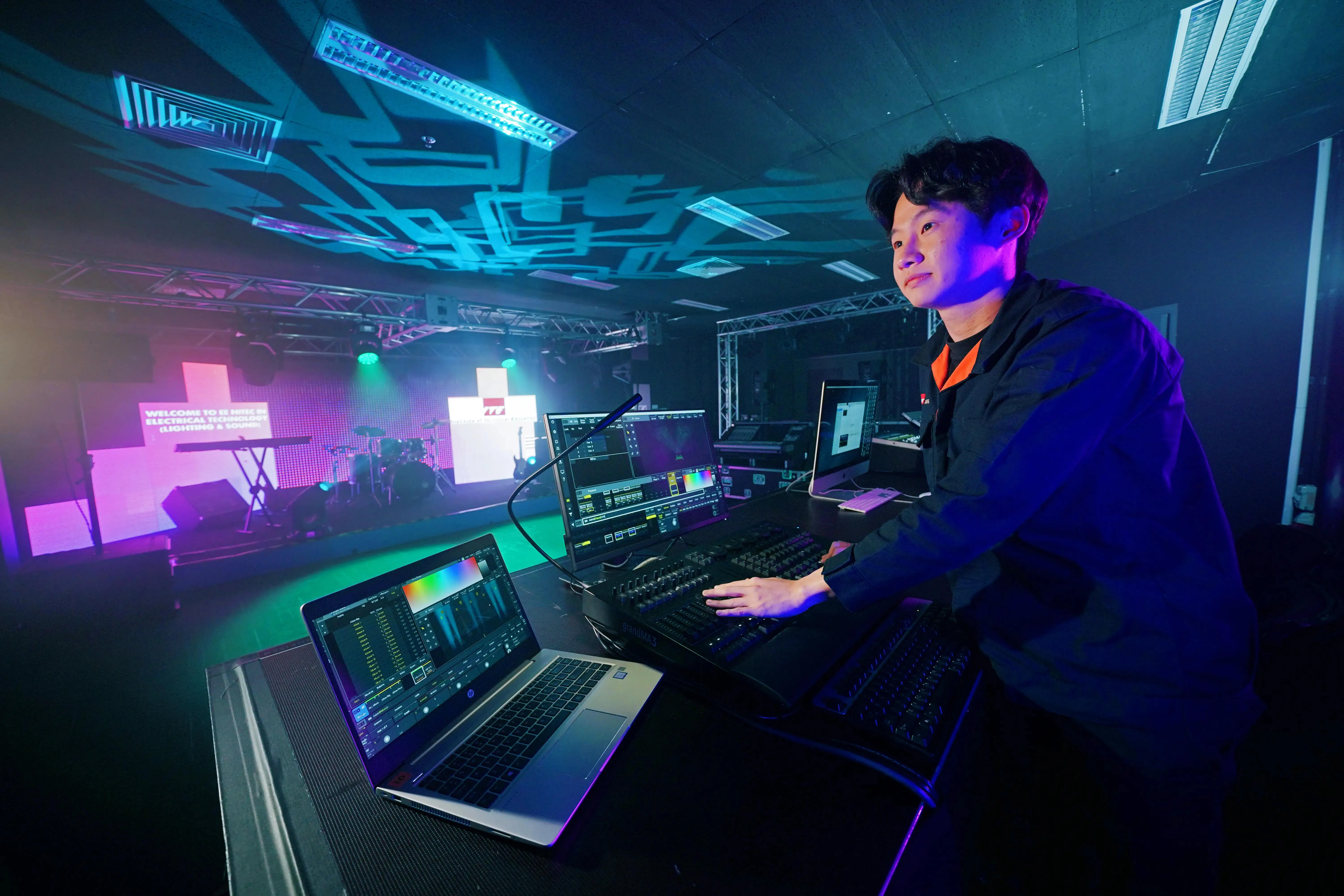 Technician using a grandMA3 lighting console, with a laptop and stage visible.