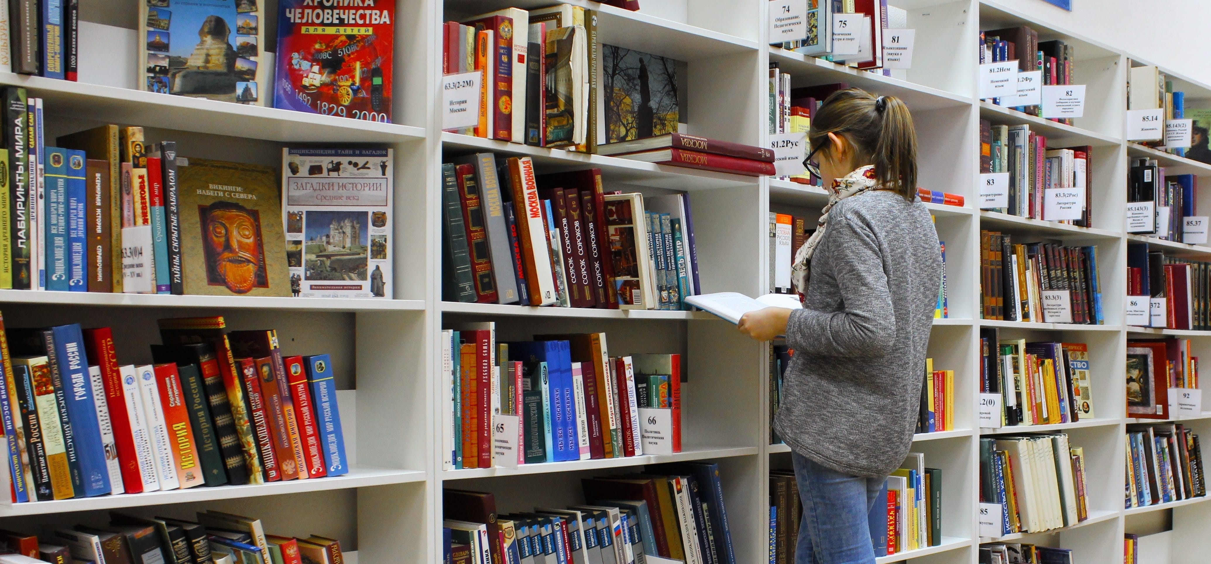 Person reading book in a library with tall shelves filled with books.