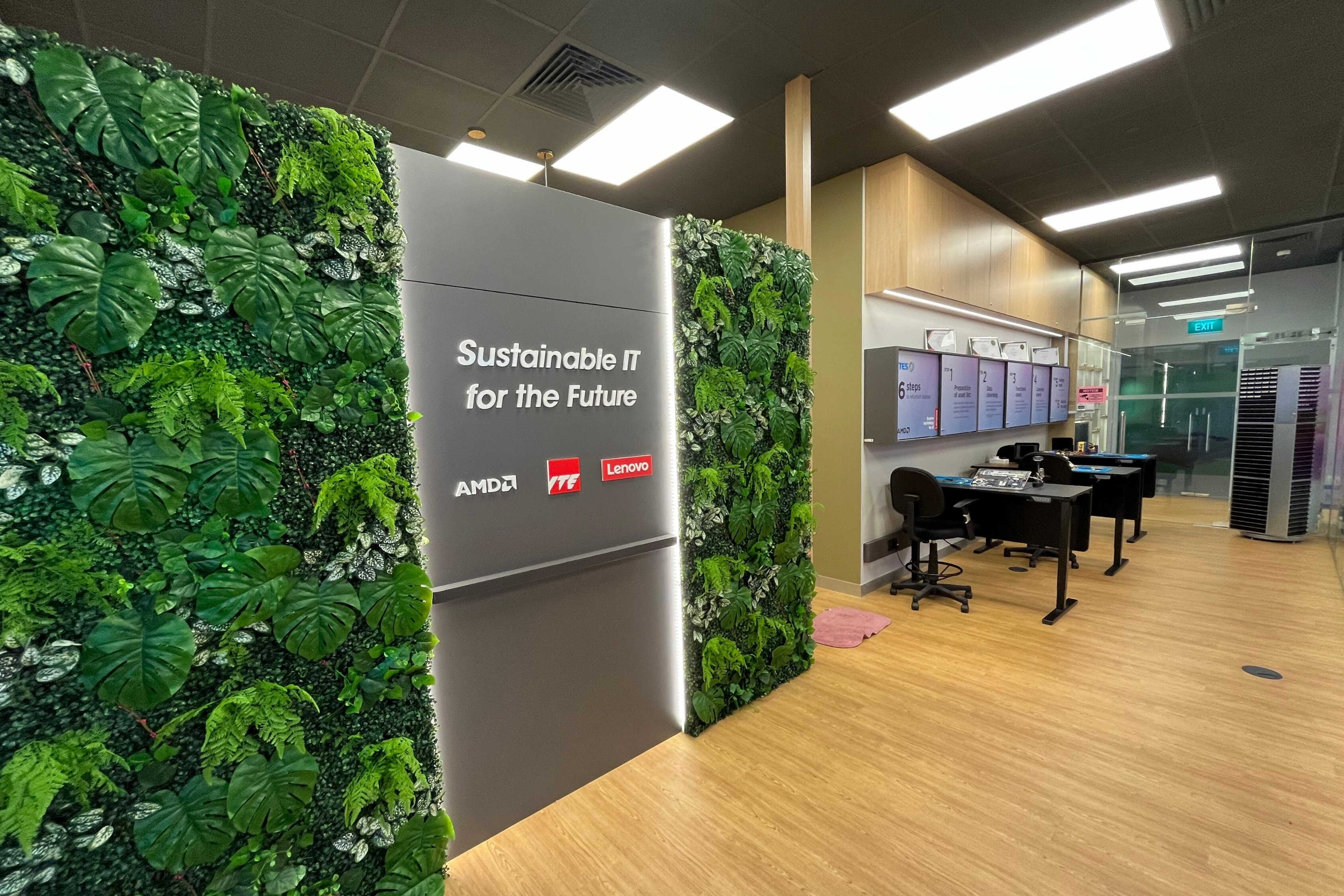Hallway with desks, foliage walls, and sign reading /'Sustainable IT for the Future/' with AMD and Lenovo logos.