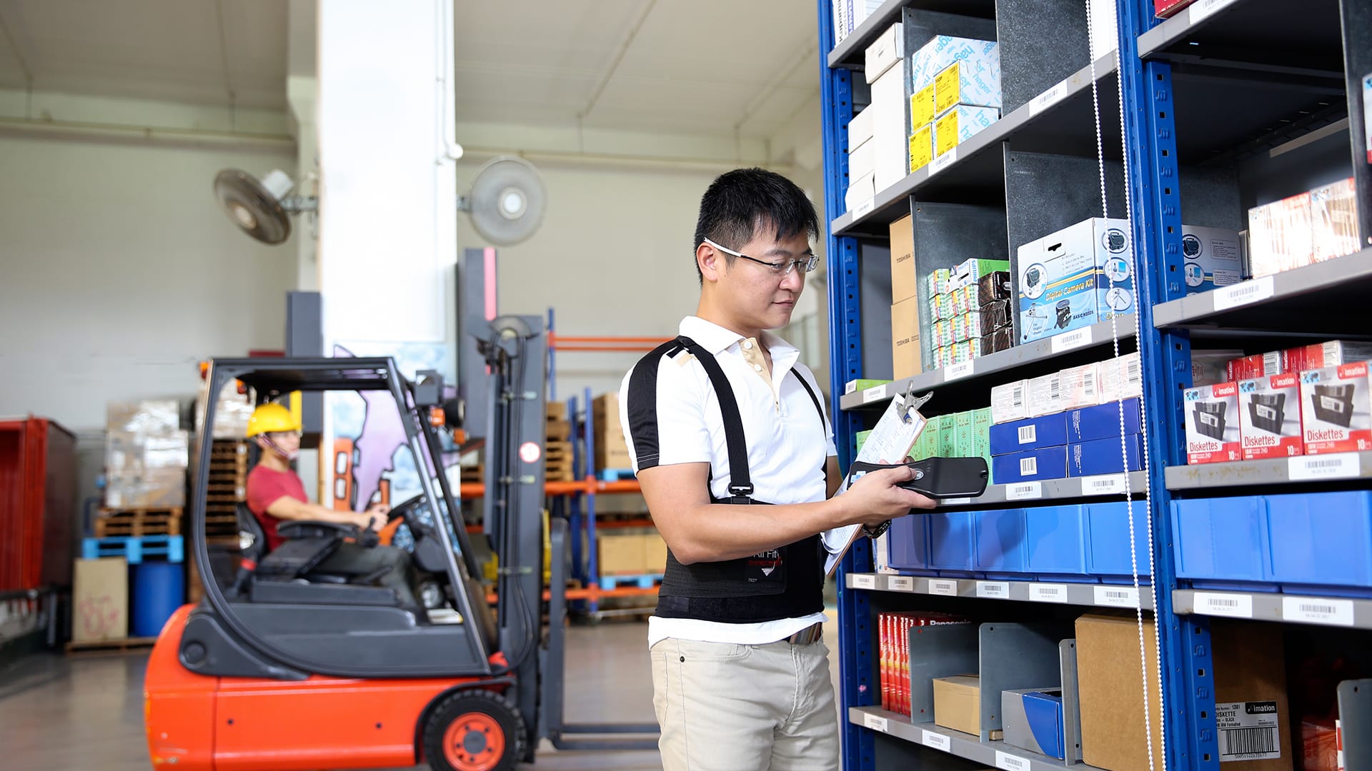 Man with glasses scanning items in warehouse with blue shelving and forklift in background.