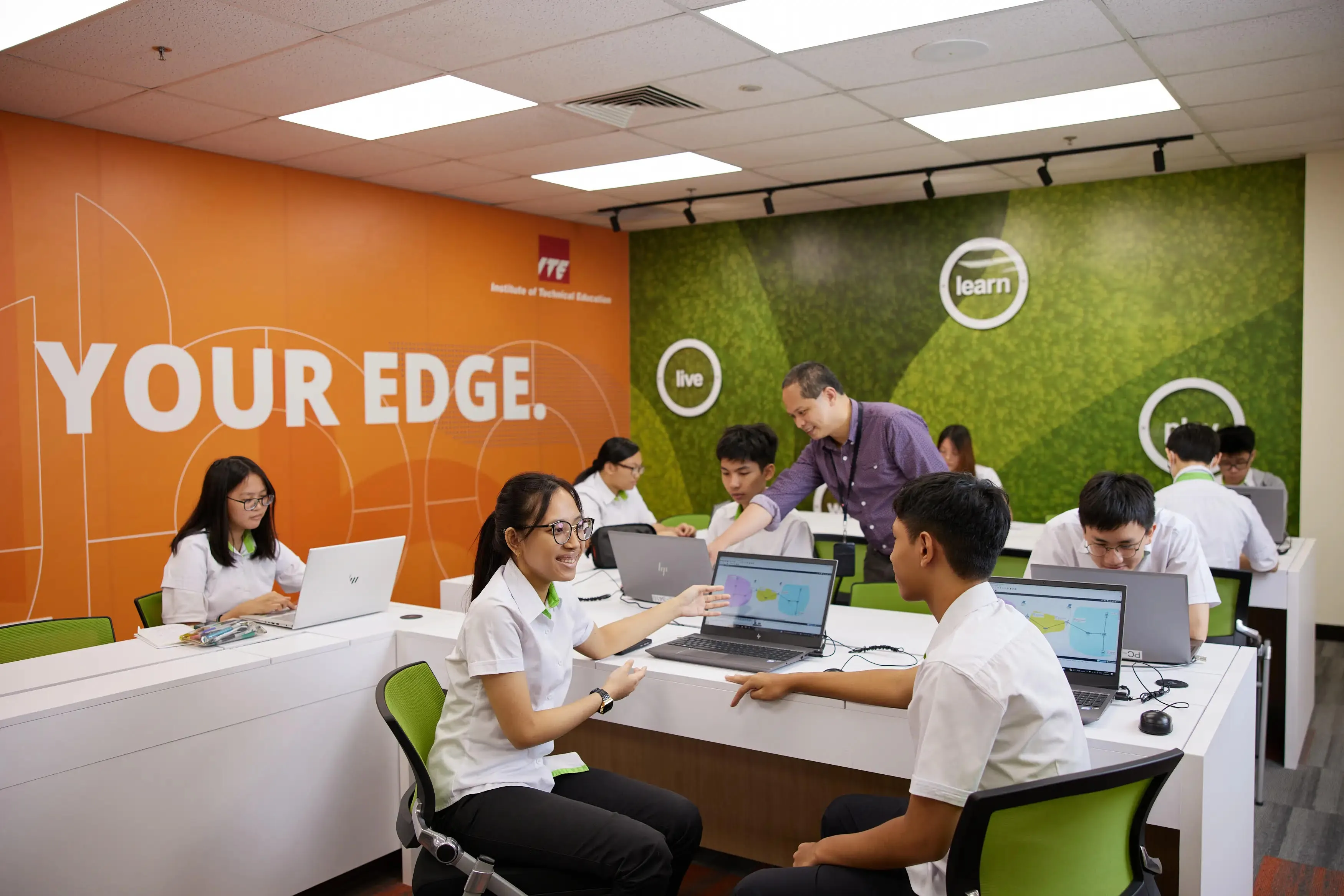 Classroom with ITE logo, students at HP laptops, teacher assisting, /'Your Edge/' and /'learn live/' on walls.
