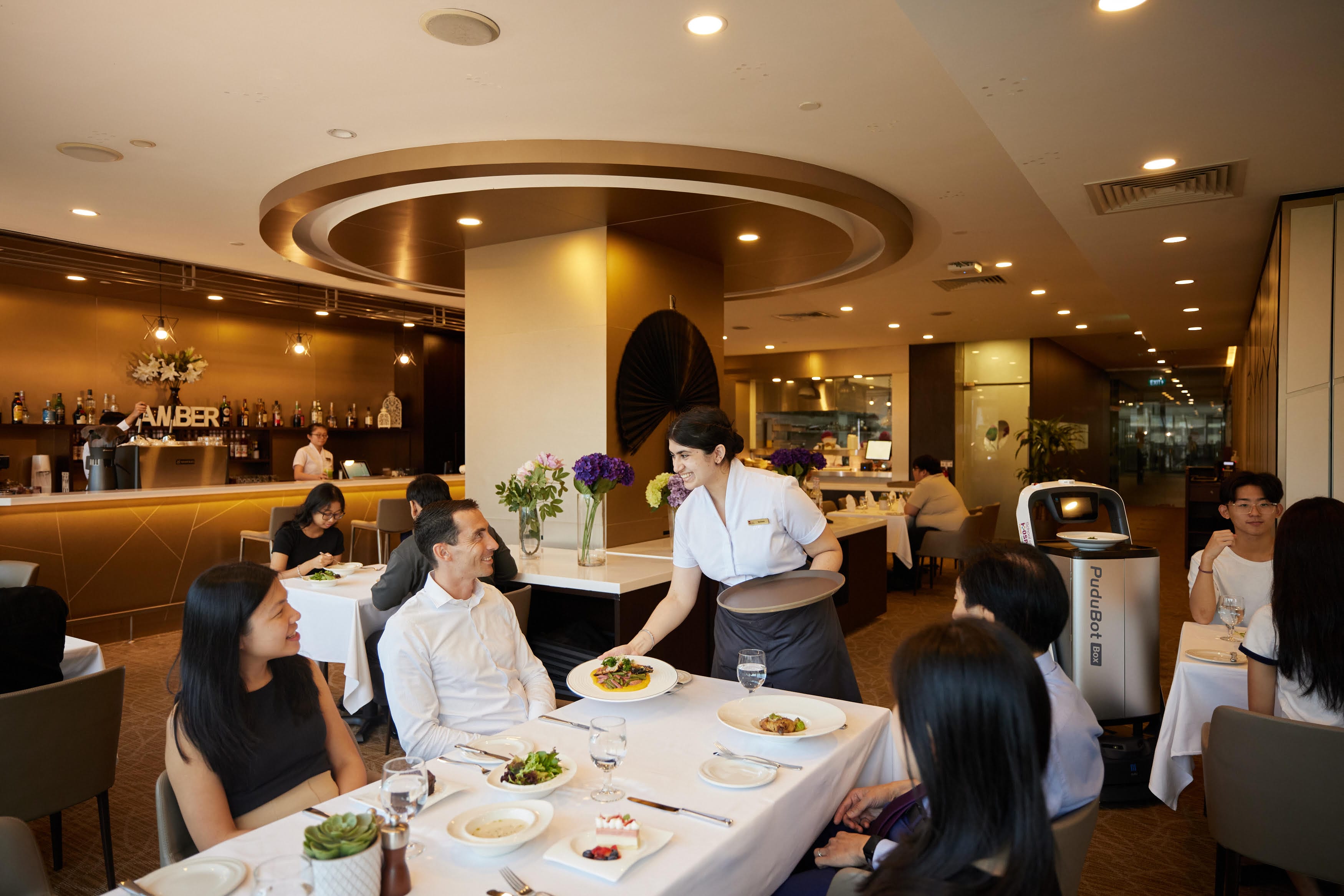 Restaurant interior; server placing dish at table, Pudubot visible.