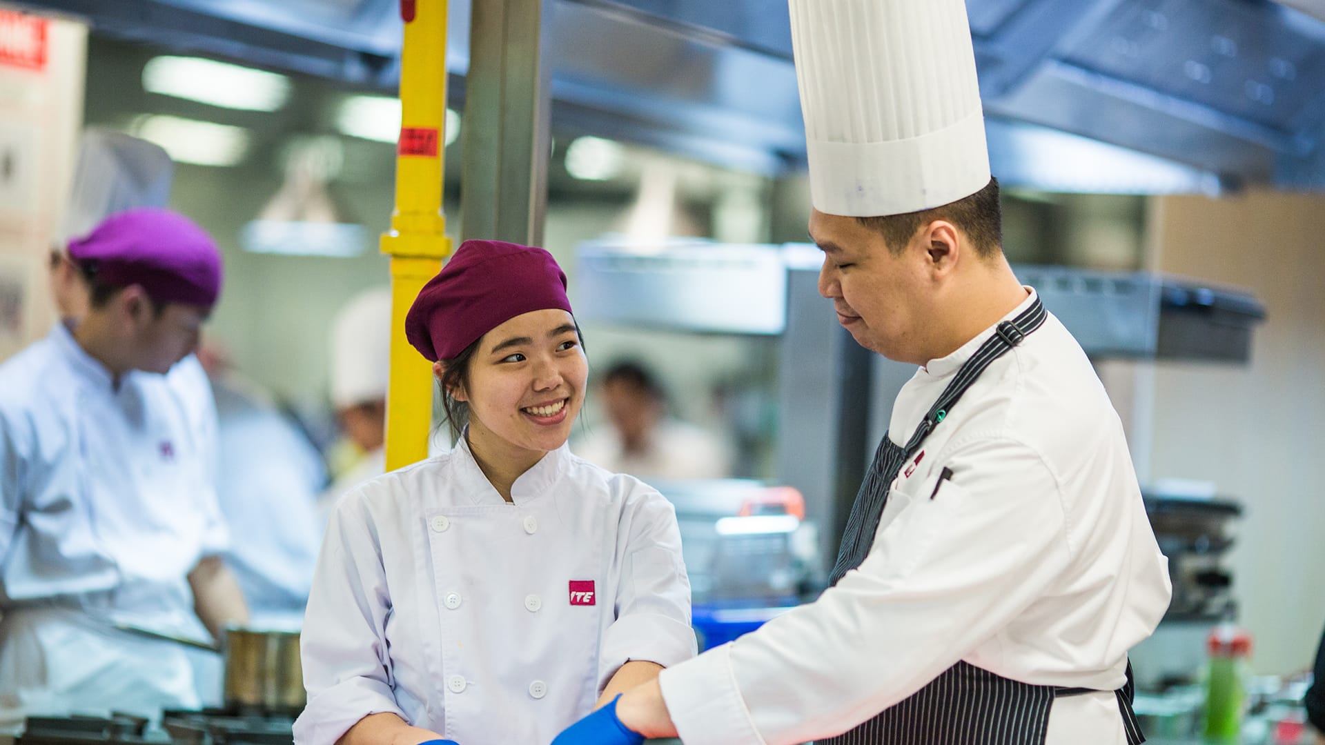 Chef guiding student in kitchen; both wearing white uniforms and chef hats