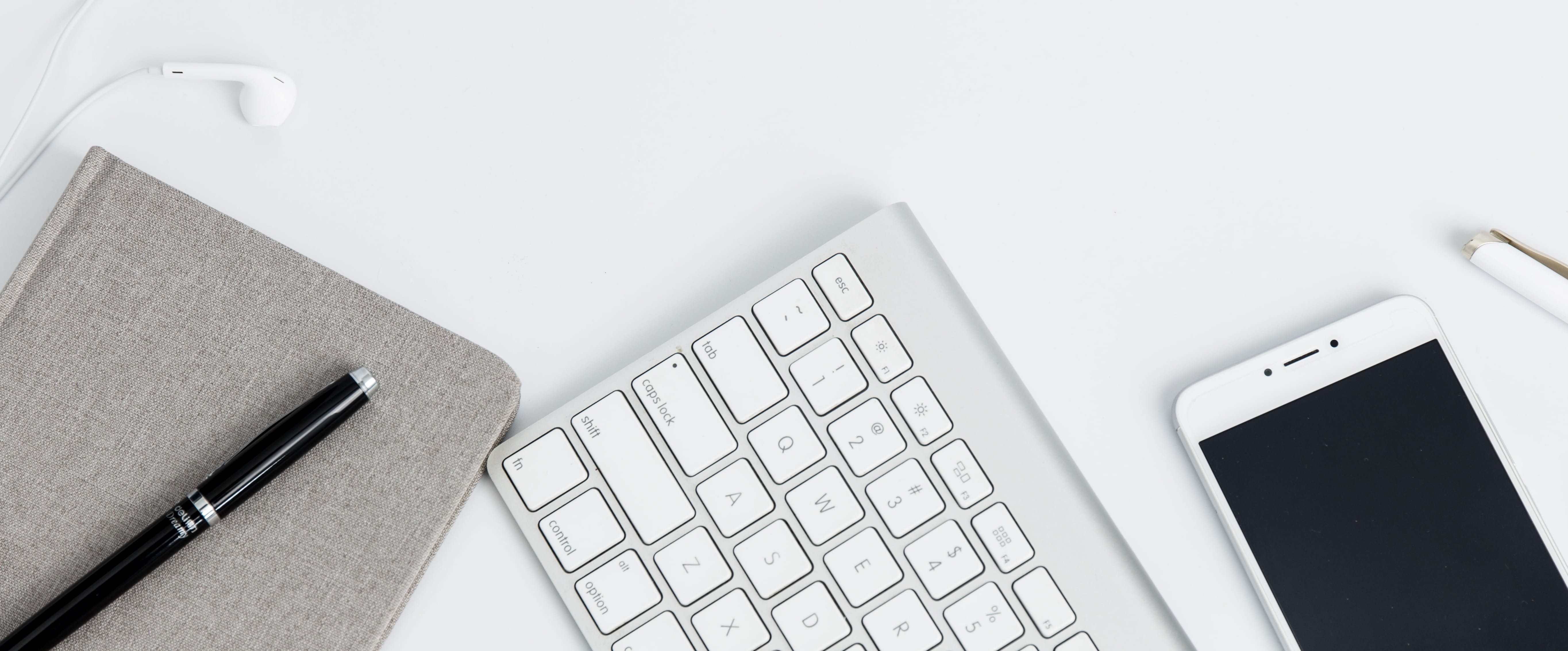 Gray notepad, black pen, Apple keyboard, white smartphone, and earbuds on a white surface.