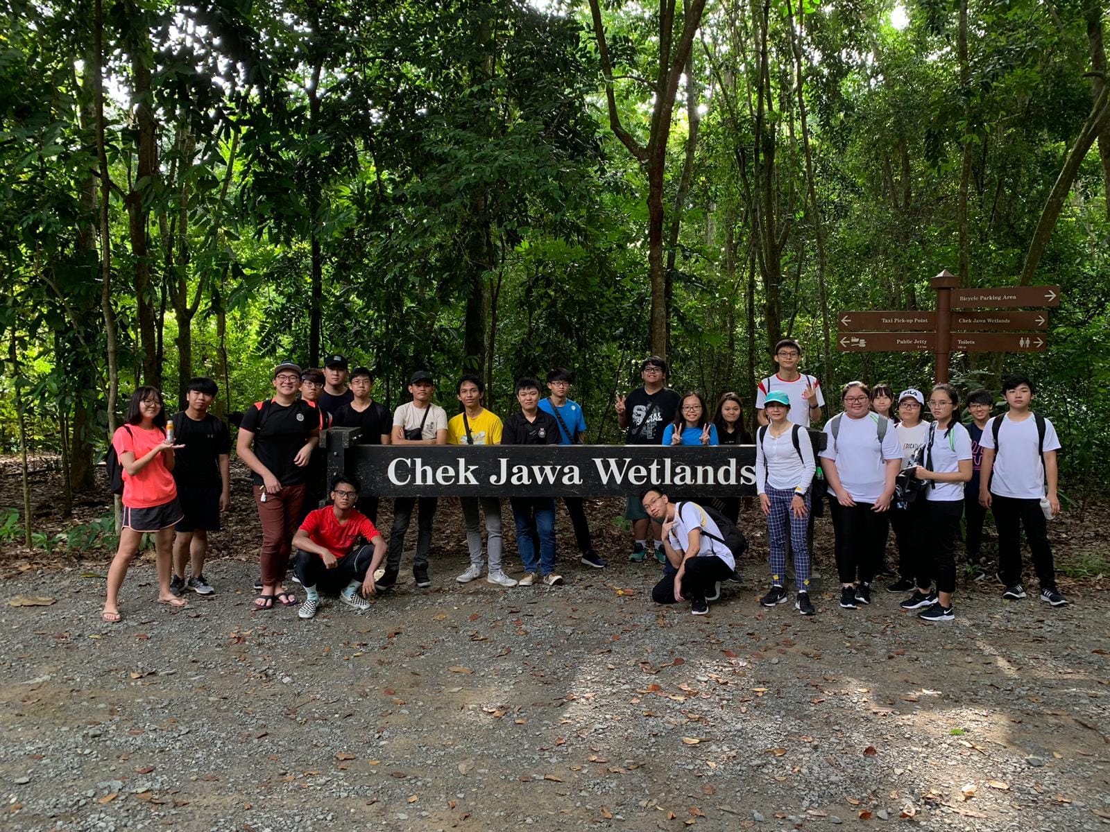Group of teens posing at 'Chek Jawa Wetlands' sign, with forest background and directional signage.