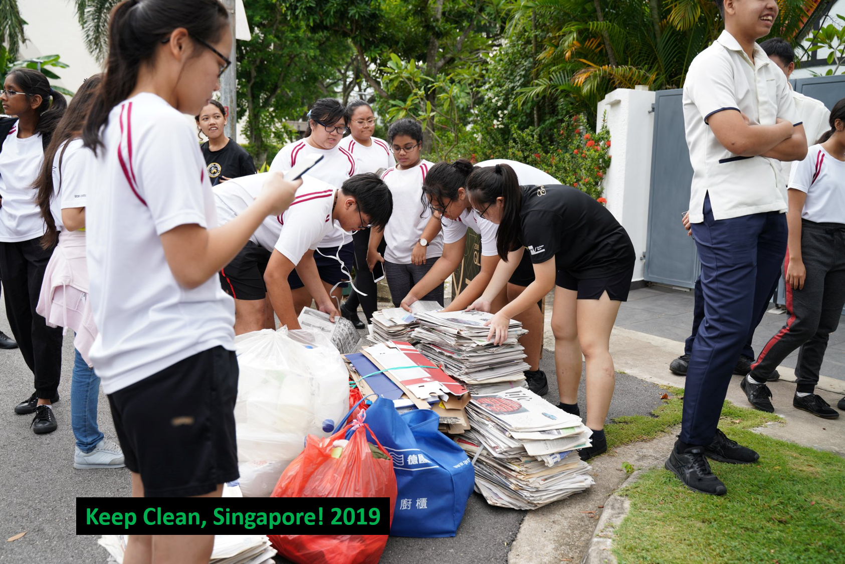 Students sort stacks of newspapers and recyclables into bags outdoors, likely for a community service project.
