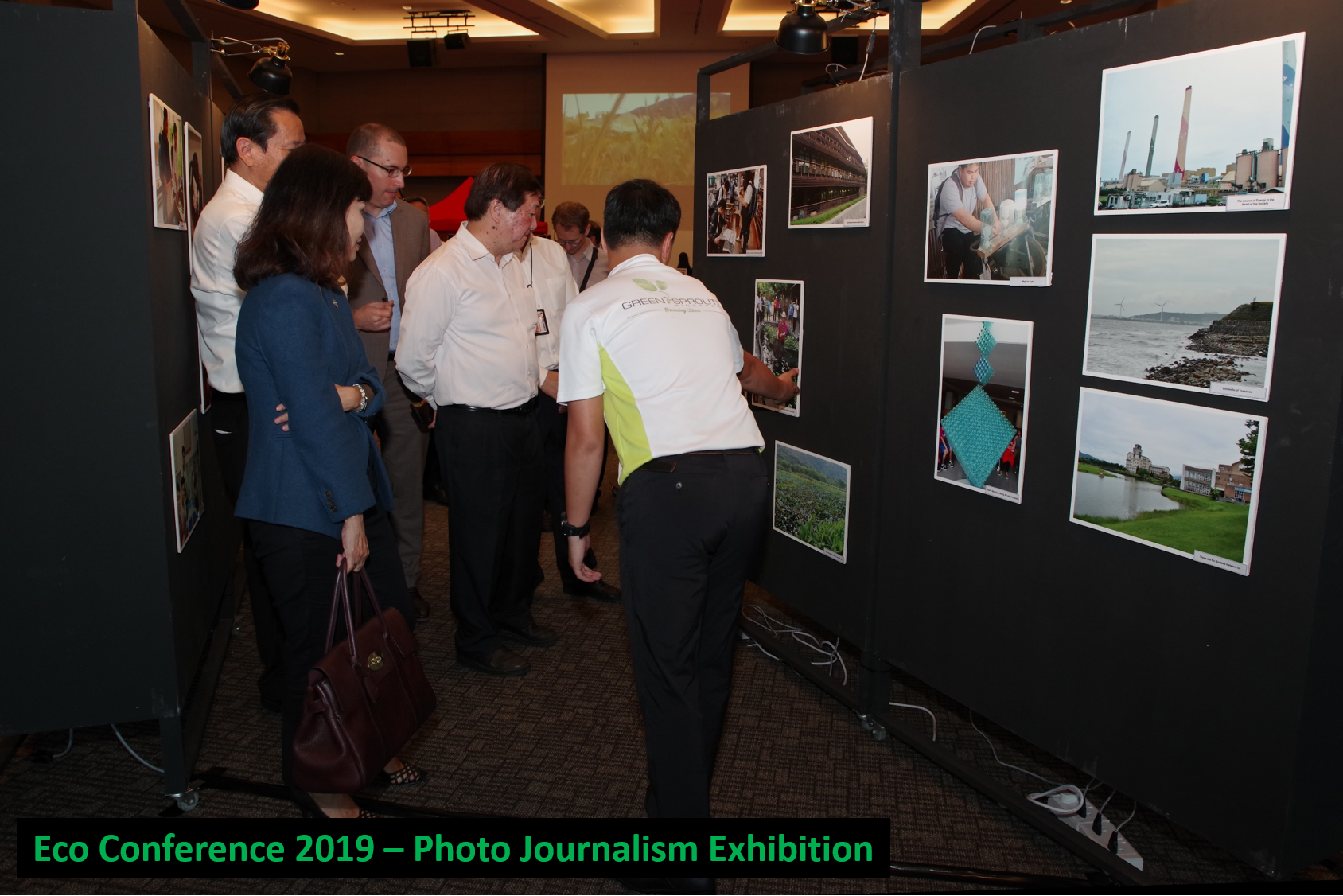 Attendees viewing a photo exhibition on black display boards in an indoor conference setting.