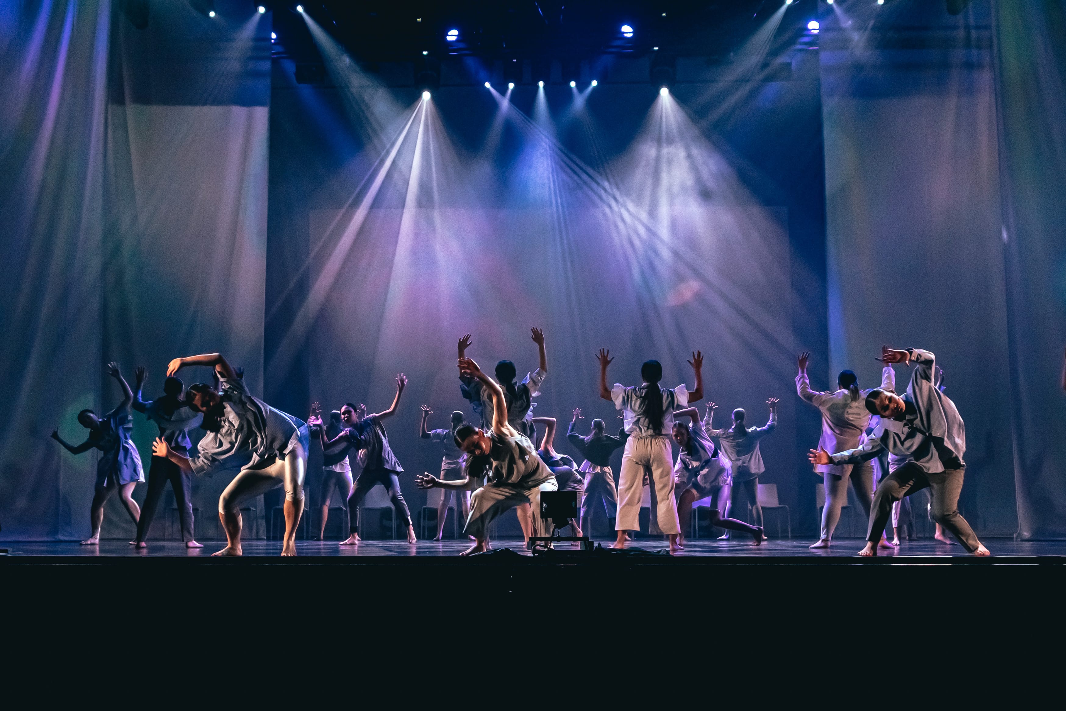 Dancers barefoot on stage in white costumes with arms raised, lit by spotlights.