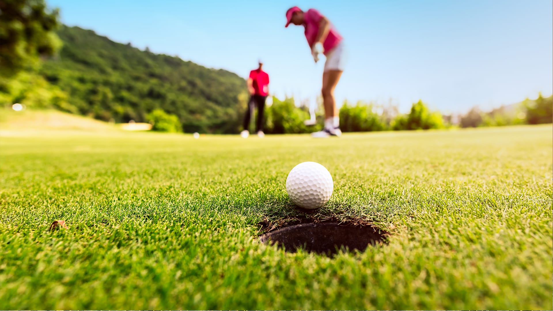 Golf ball at the edge of a hole on the green, golfers blurred in the background.