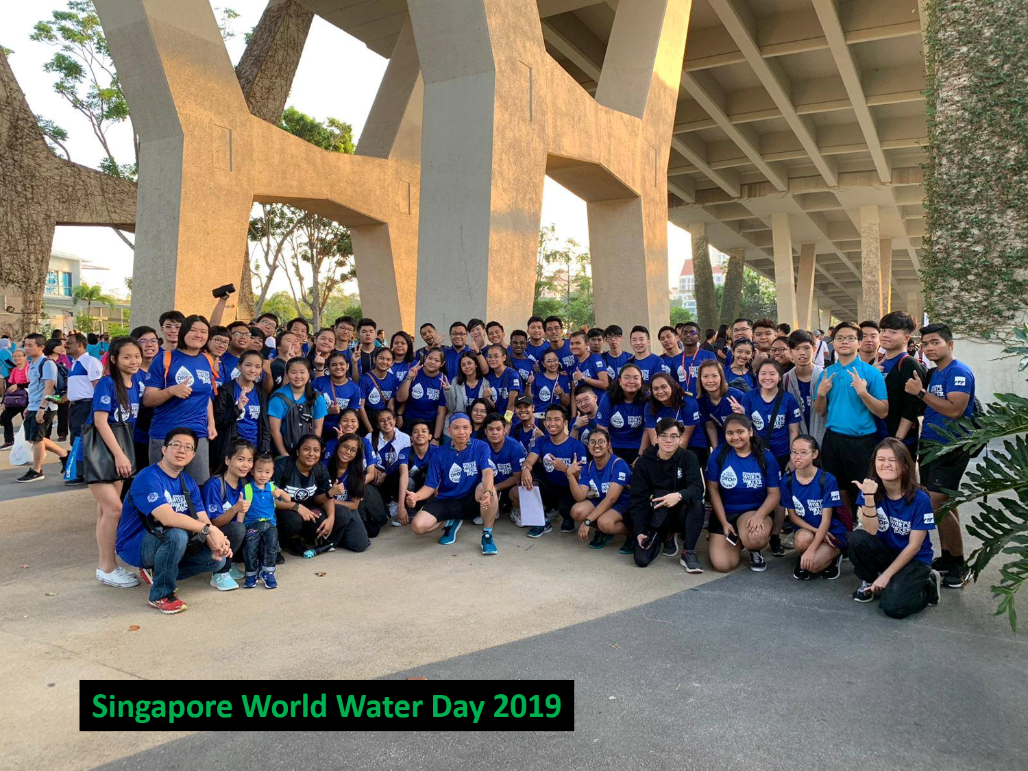 Large group wearing blue shirts, posing outdoors under a large concrete structure.