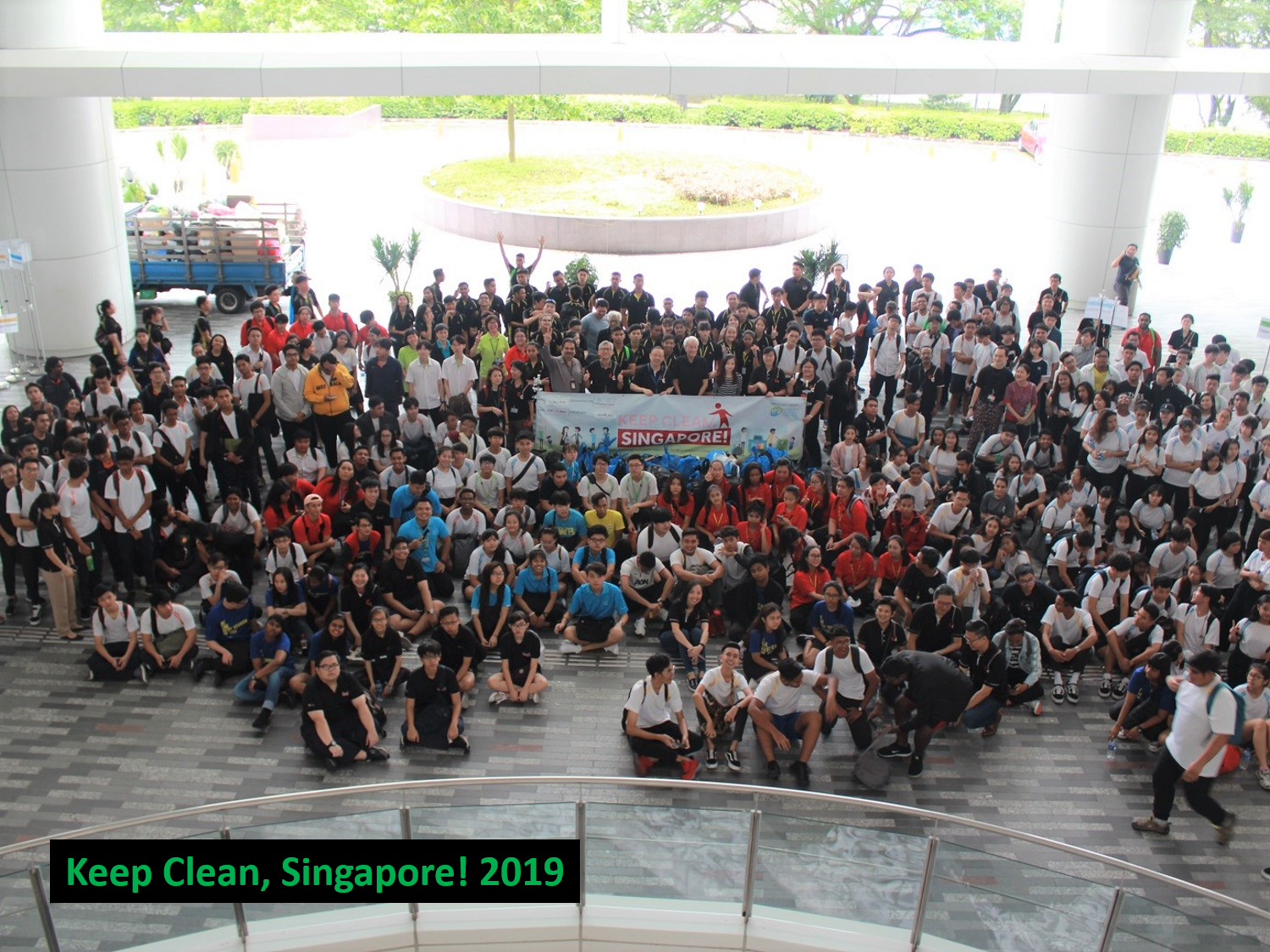 Large group of people pose with 'Keep Clean, Singapore!' banner in a plaza.