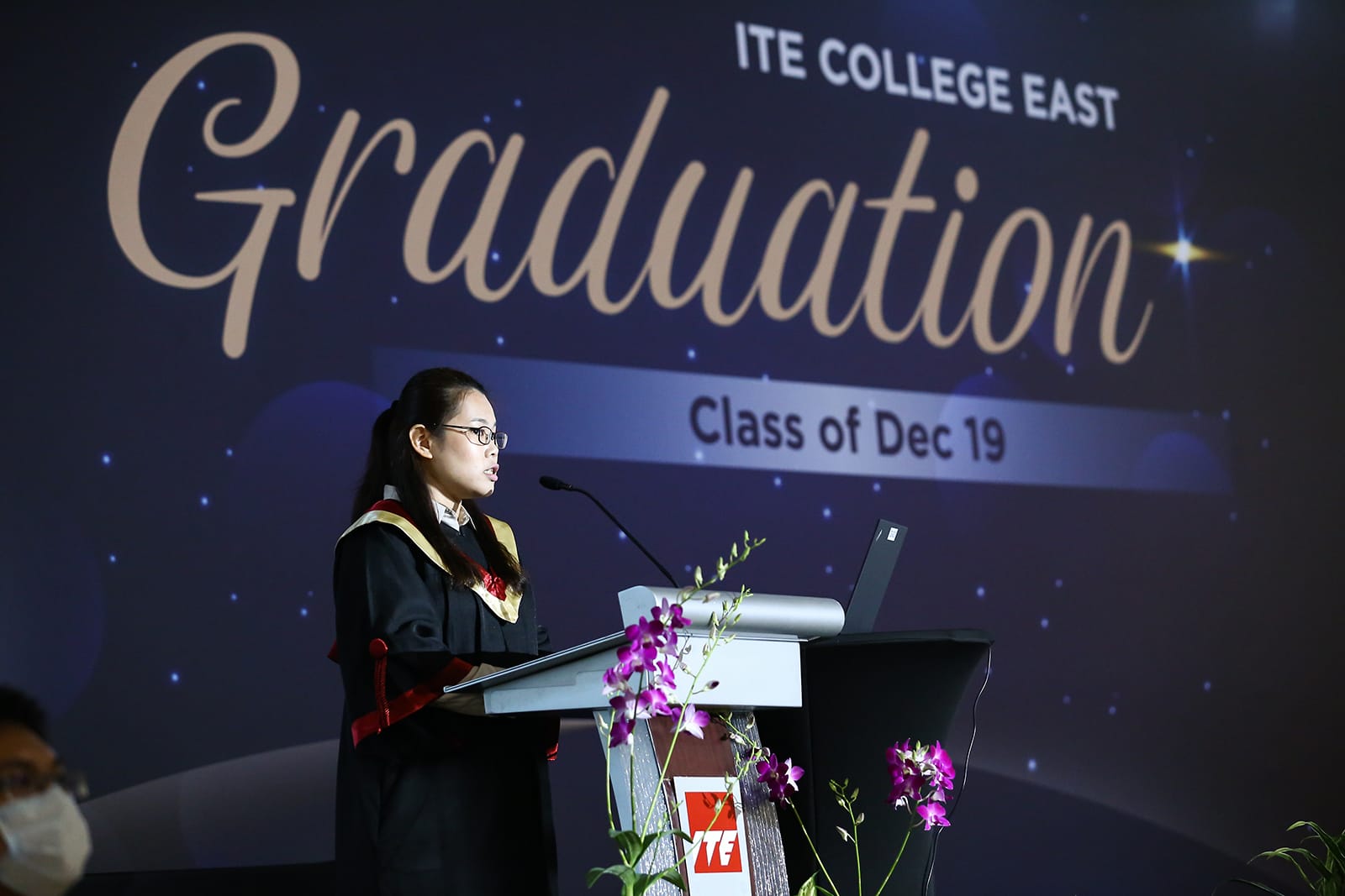 Woman in graduation gown at podium speaking, ITE College East backdrop.