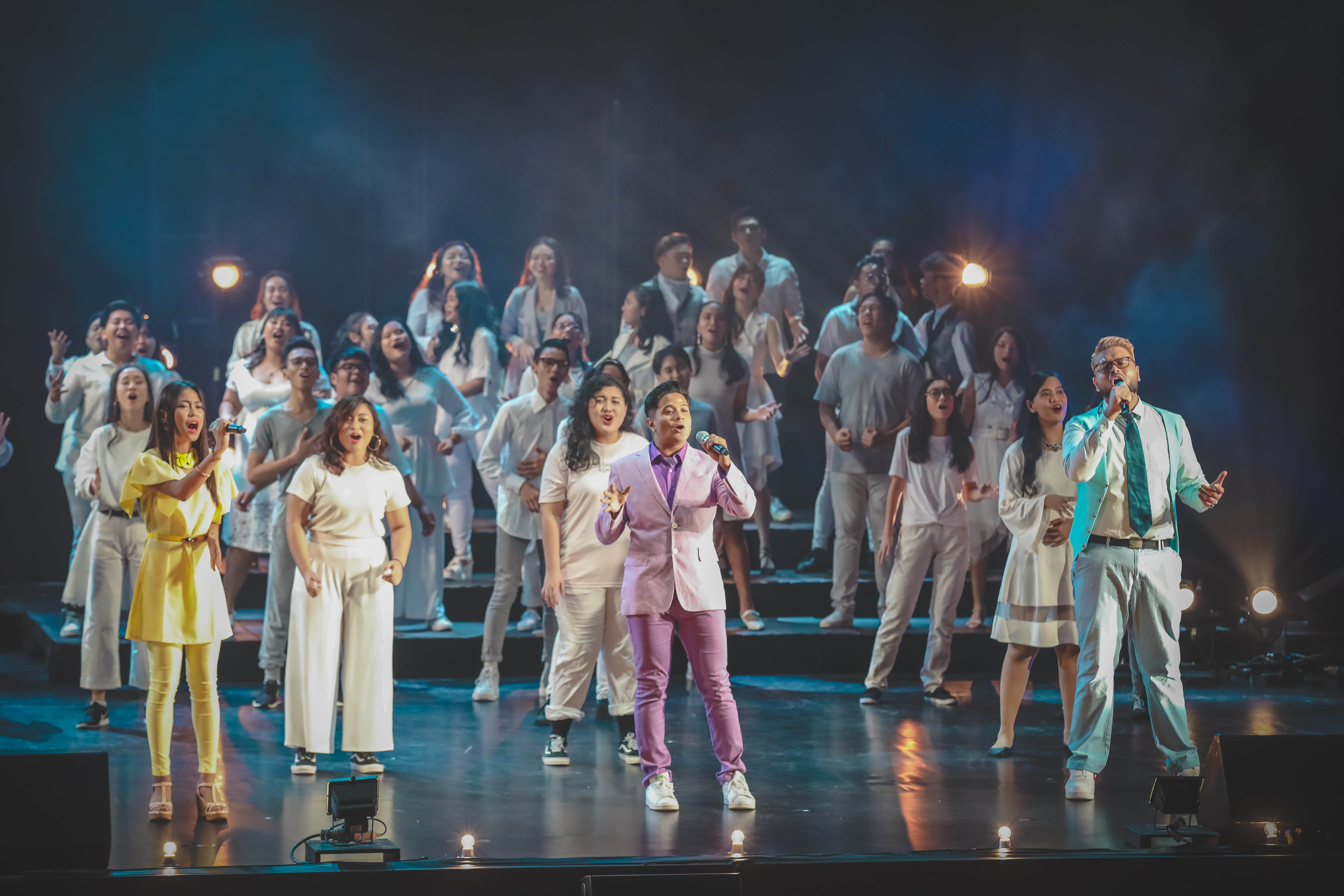 Choir on stage: singers in white, leaders in lilac and teal, performing with microphones.
