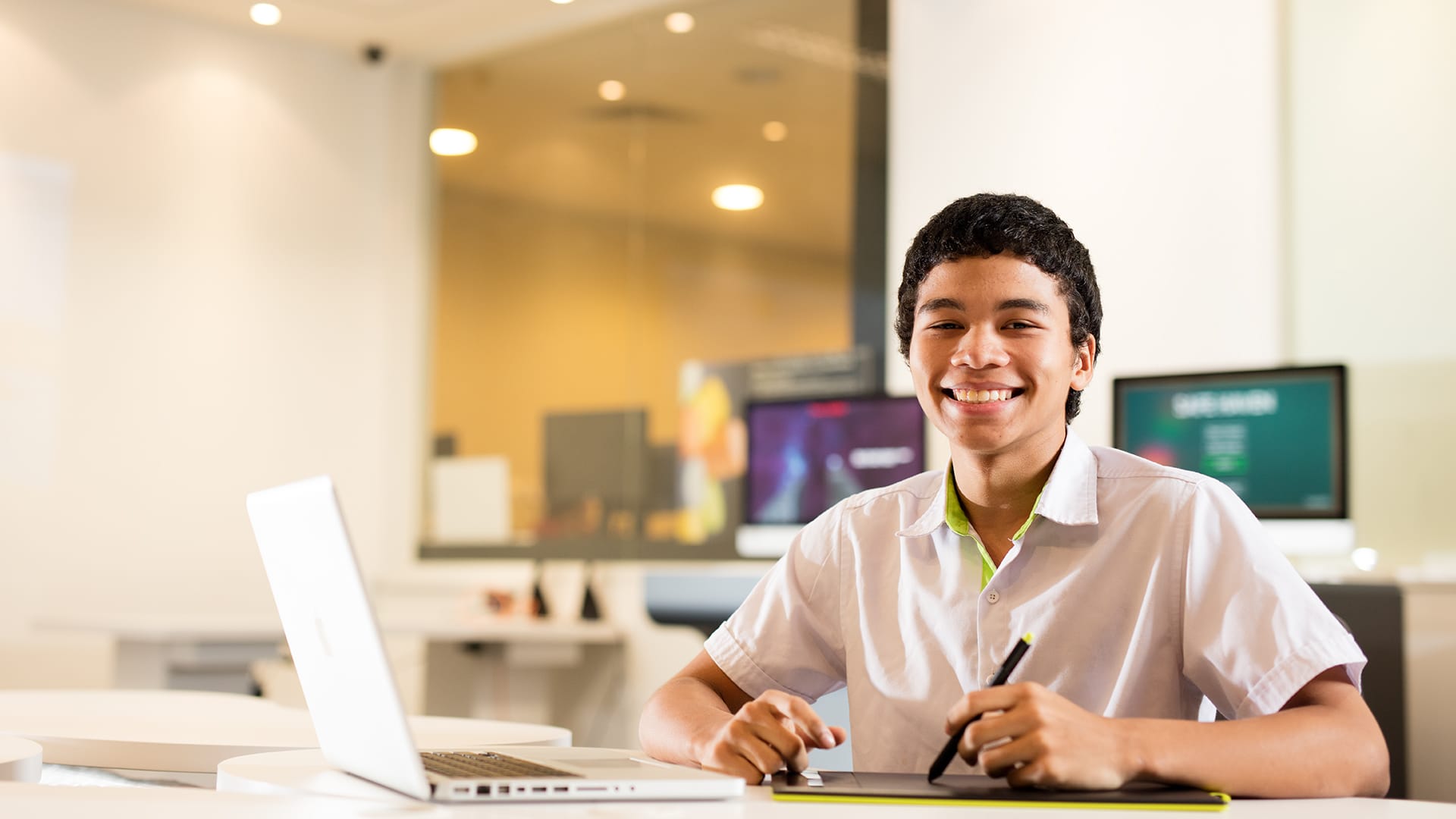 Student smiling at laptop in a bright classroom with blurred monitors behind