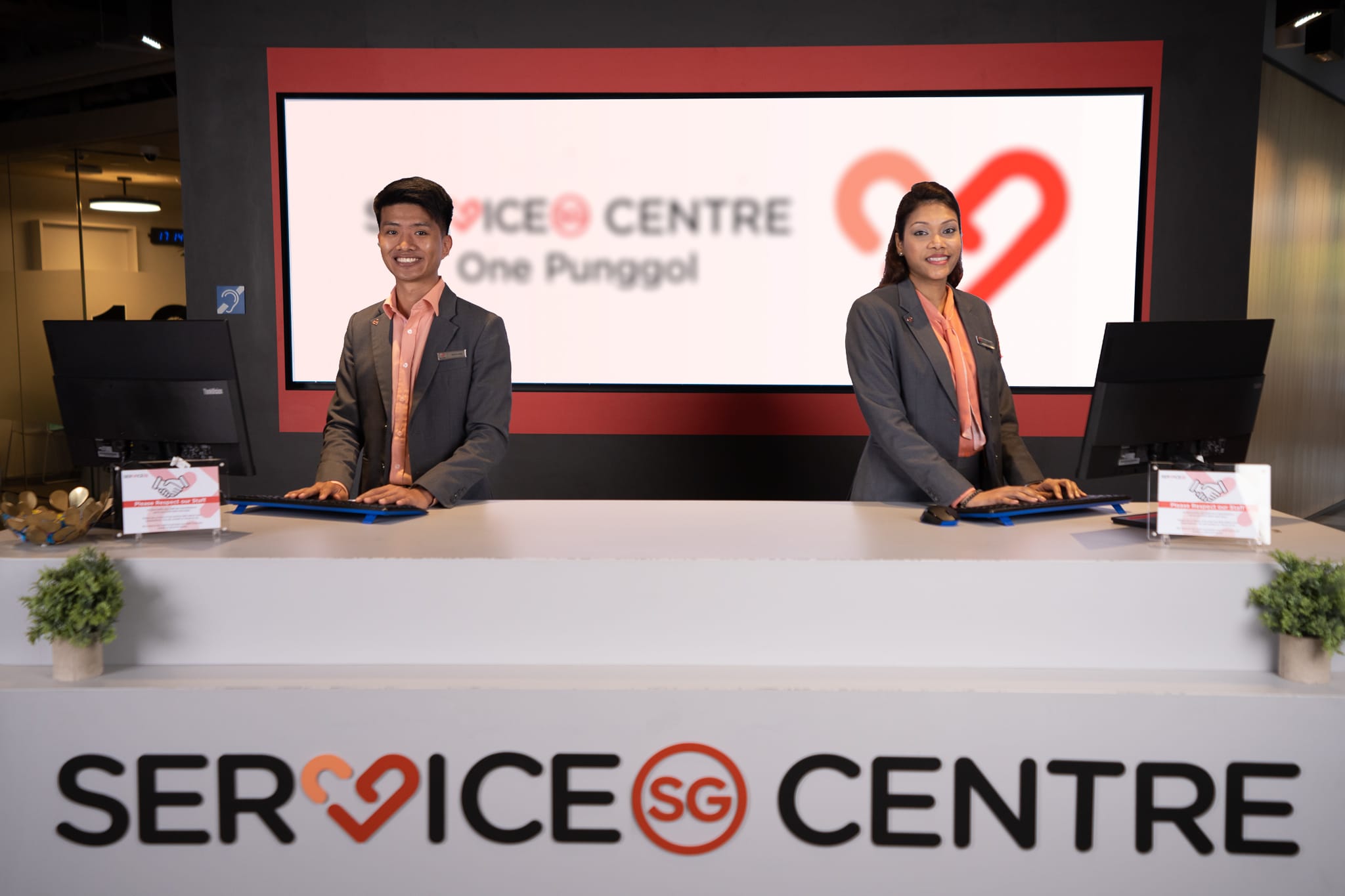 Two ServiceSG officers at the One Punggol Centre front desk with logo in background