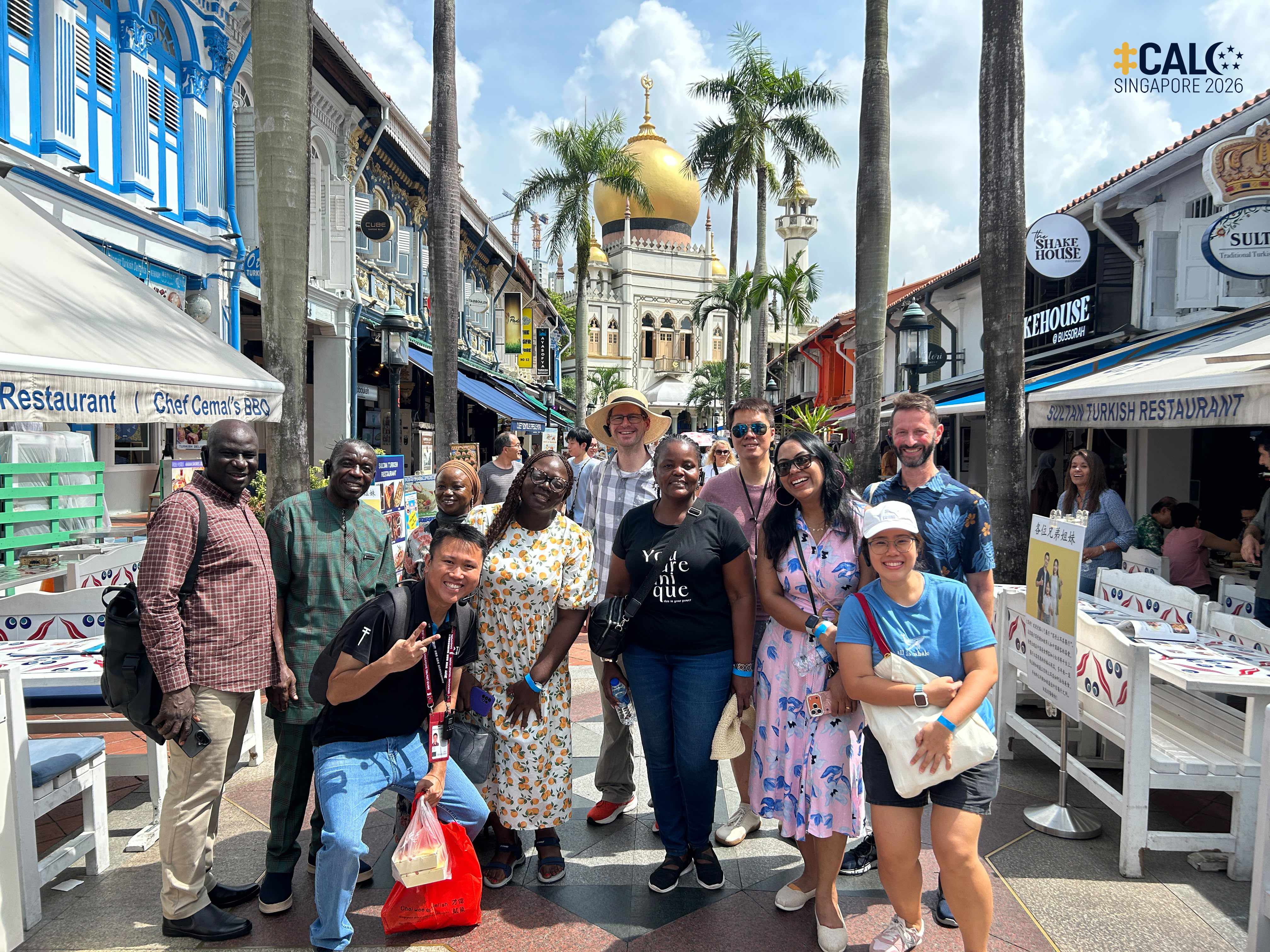Conference delegates pose for a group photo along a pedestrianised street in Kampong Gelam, with shophouses lining both sides of the street and Sultan Mosque in the background, on 18 July 2026.