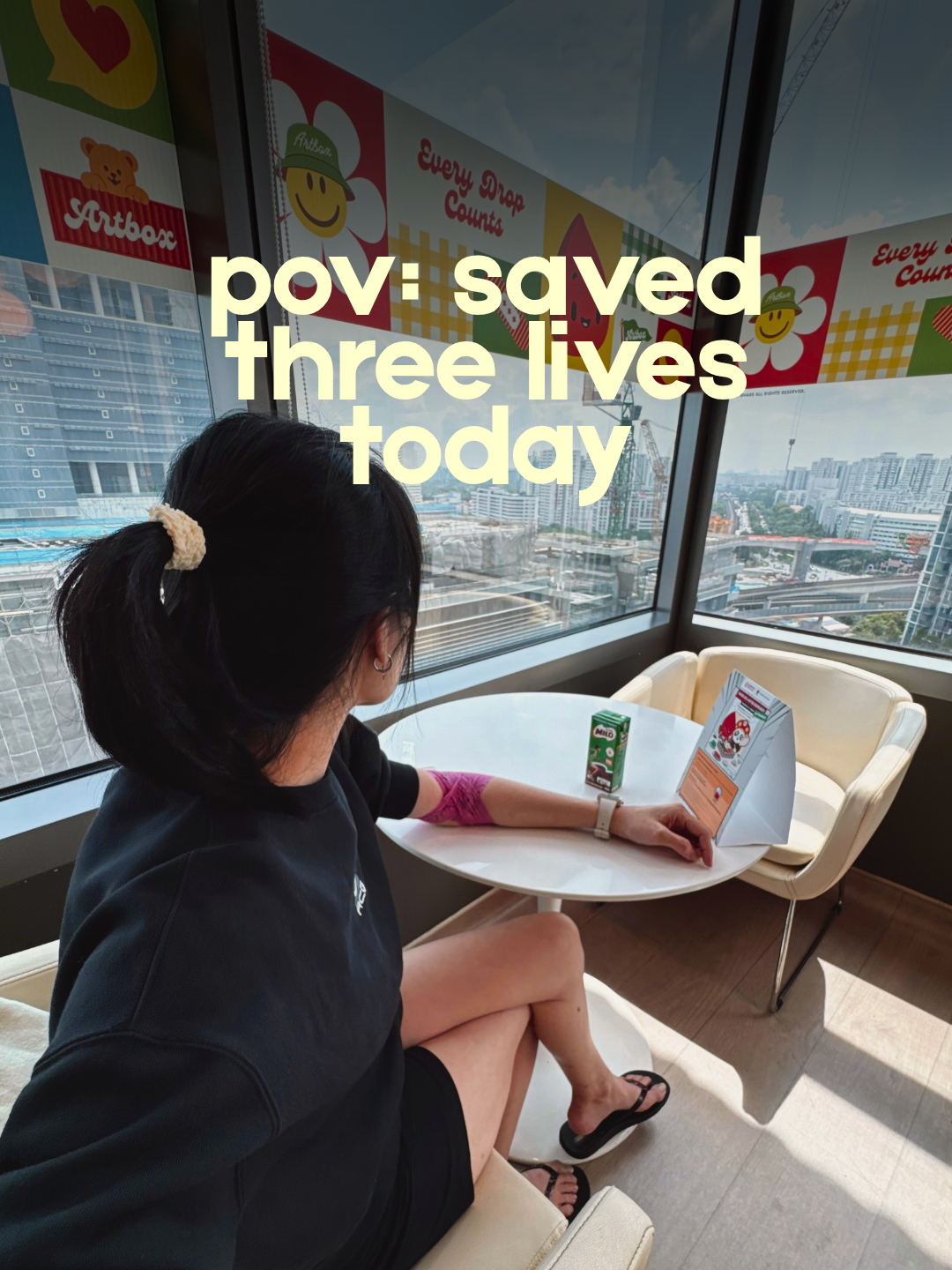 A woman with a pink bandage on her arm sits at a table looking out a high-rise window overlooking a city skyline. The text overlay reads, "pov: saved three lives today."