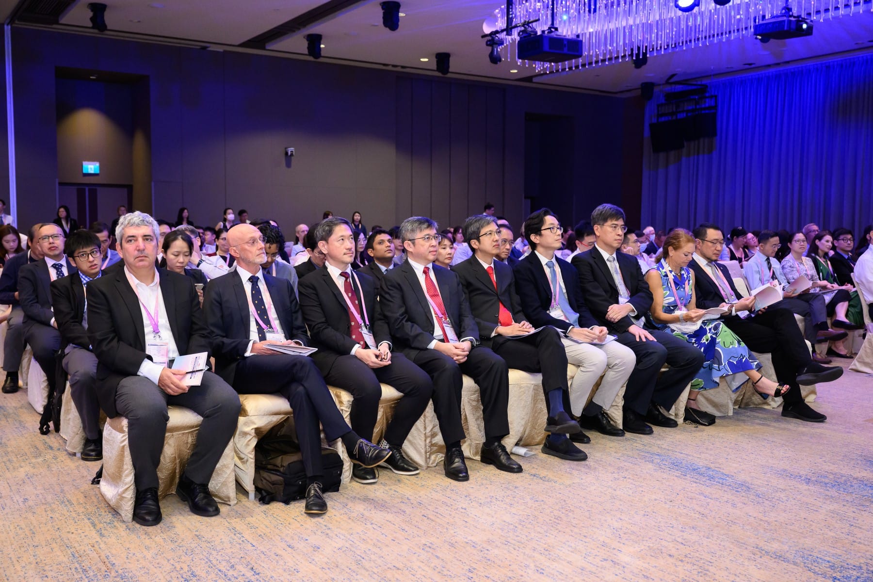 Audience of people in suits sitting in rows at conference with blue lighting.