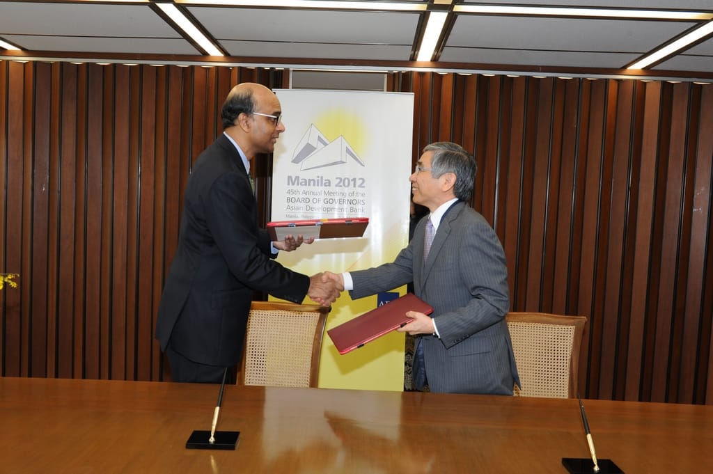 Two men in suits shake hands, each holding a red folder. Banner behind: "Manila 2012 45th Annual Meeting Asian Development Bank"