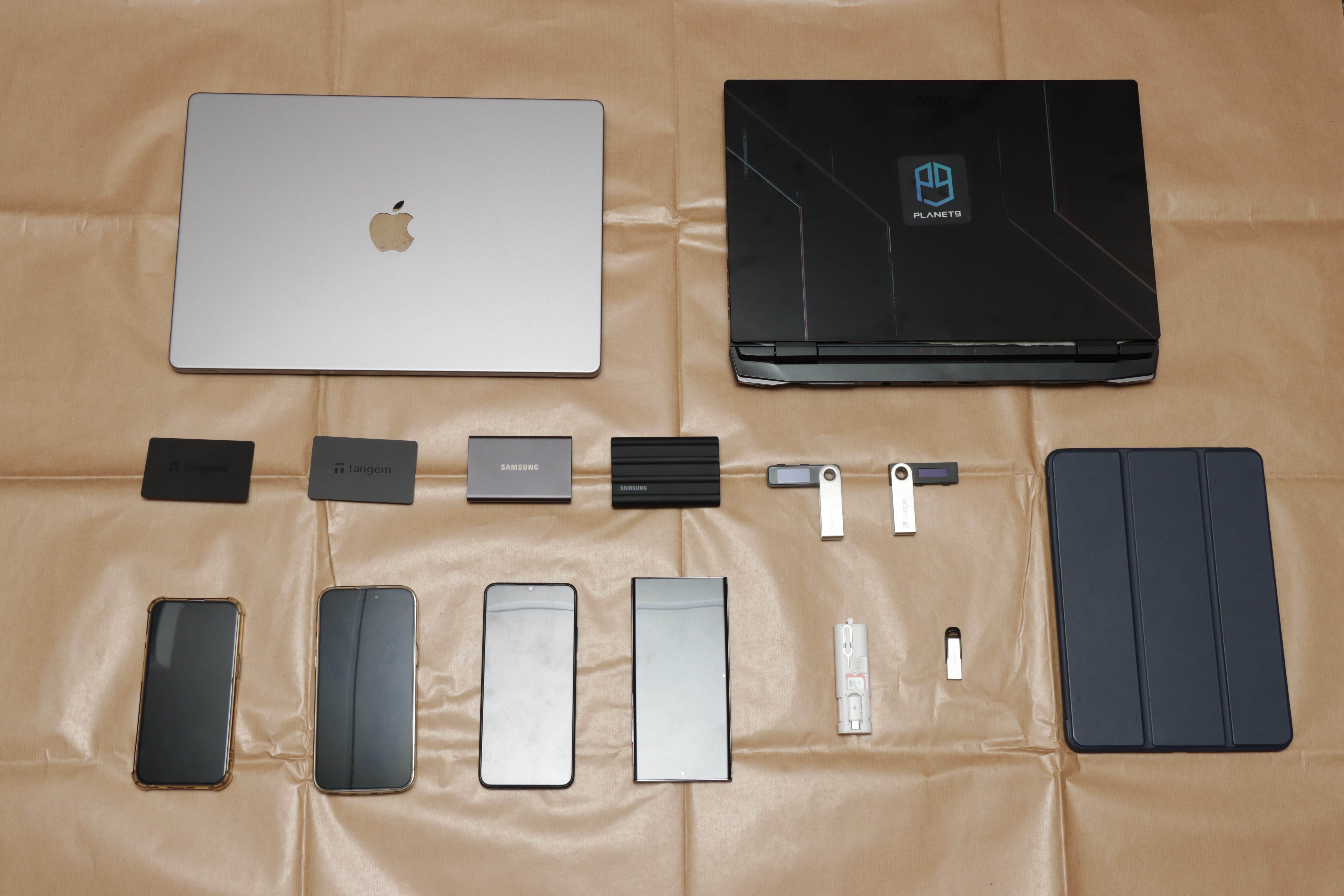 Laptops, phones, USB drives, hard disks, and crypto wallets arranged on brown paper.