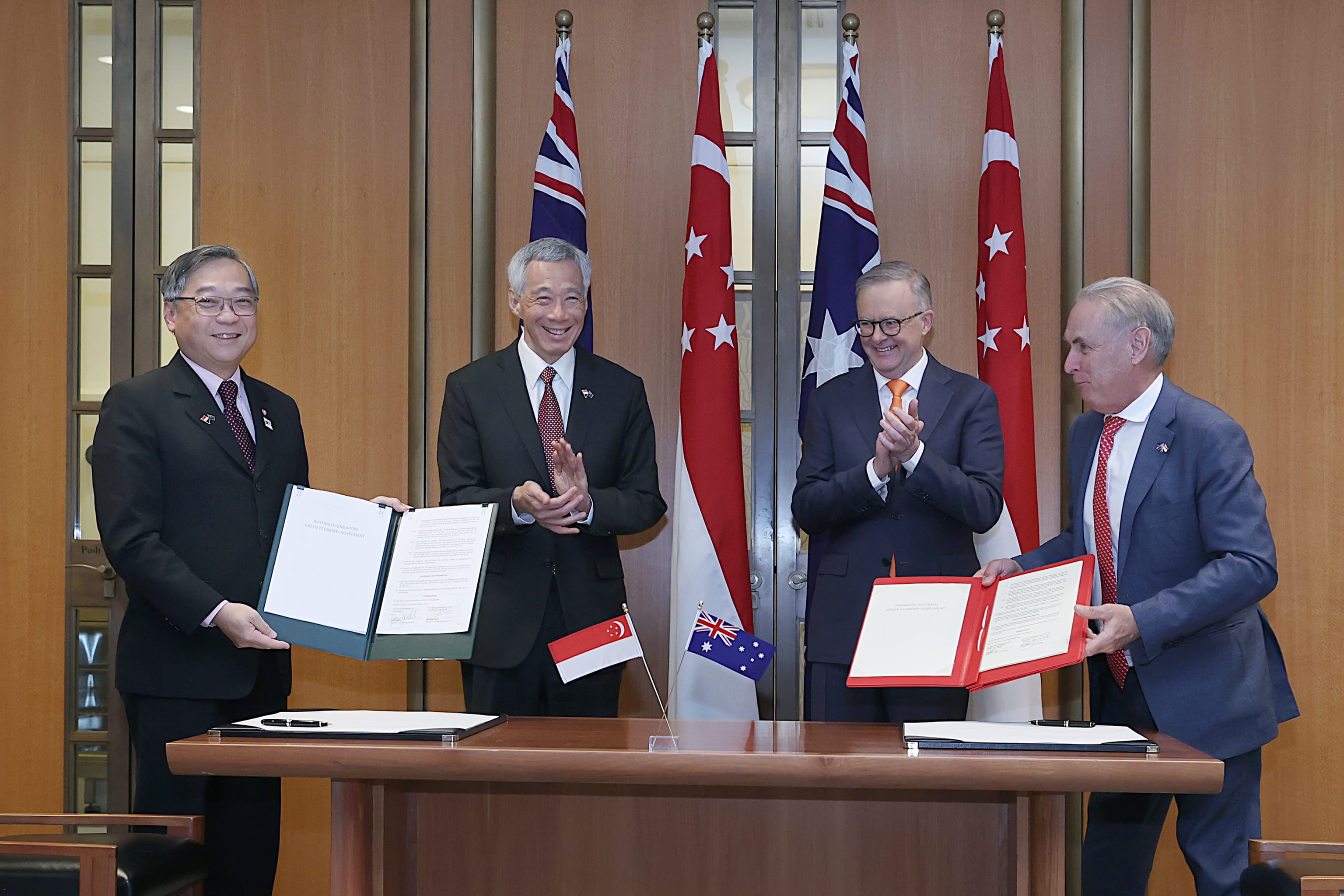 Four men in suits holding folders, standing before Australian & Singaporean flags.