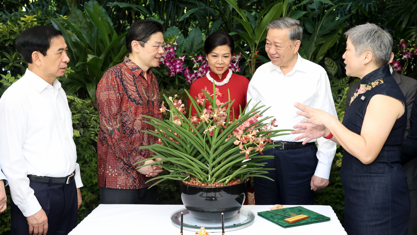 Five people surround a floral arrangement on a table, in front of green foliage.