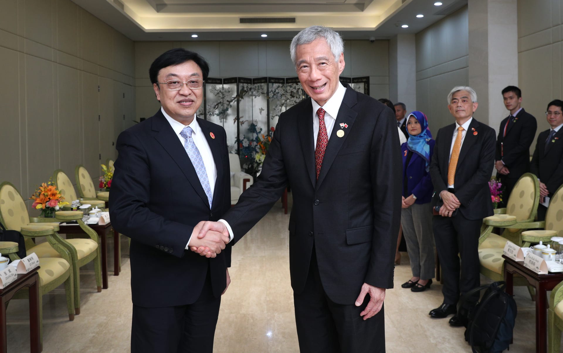 Two men in suits shake hands. Lee Hsien Loong on right. Others visible in the background.