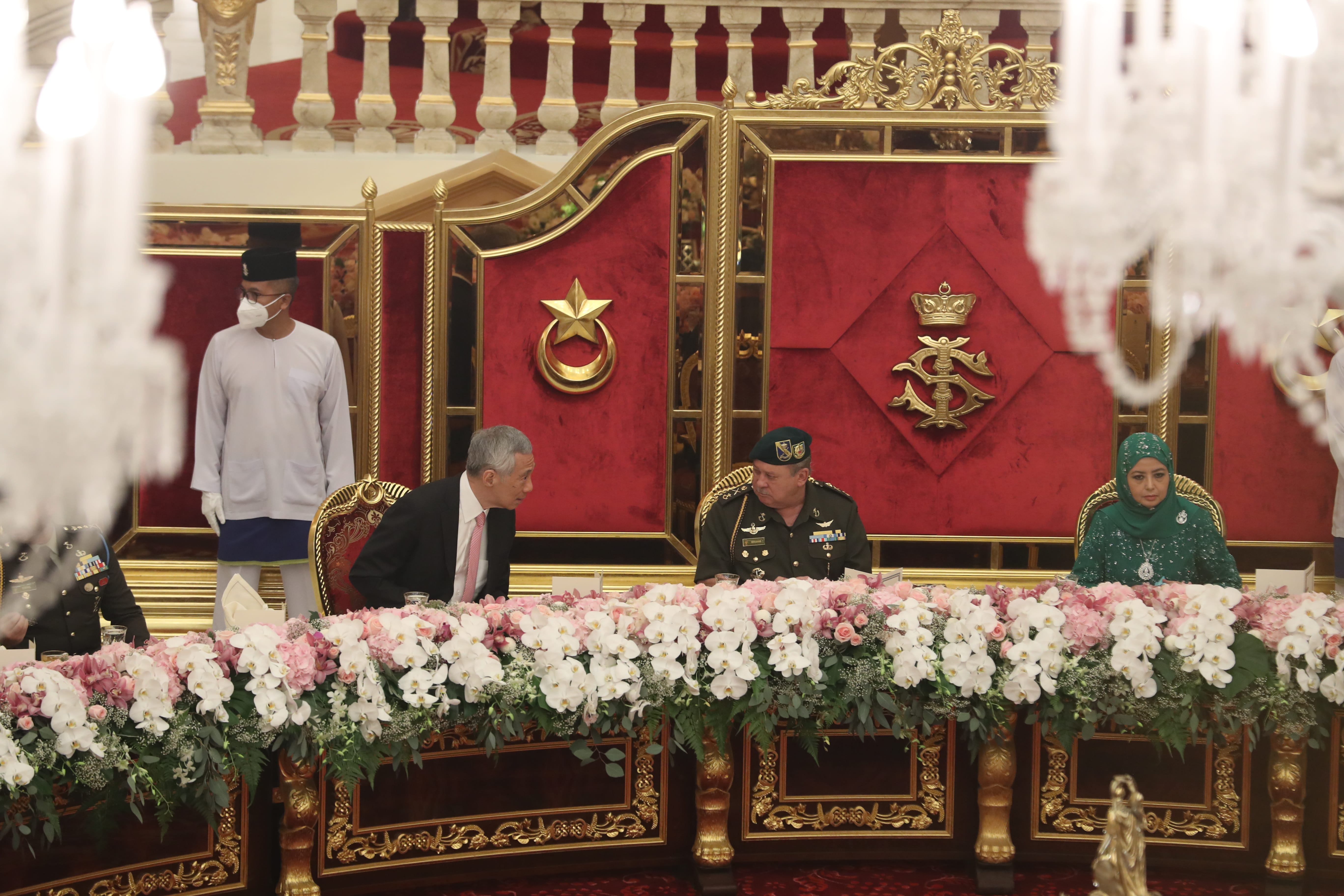 People seated behind an ornate table covered in flowers, with emblems on the wall behind them.