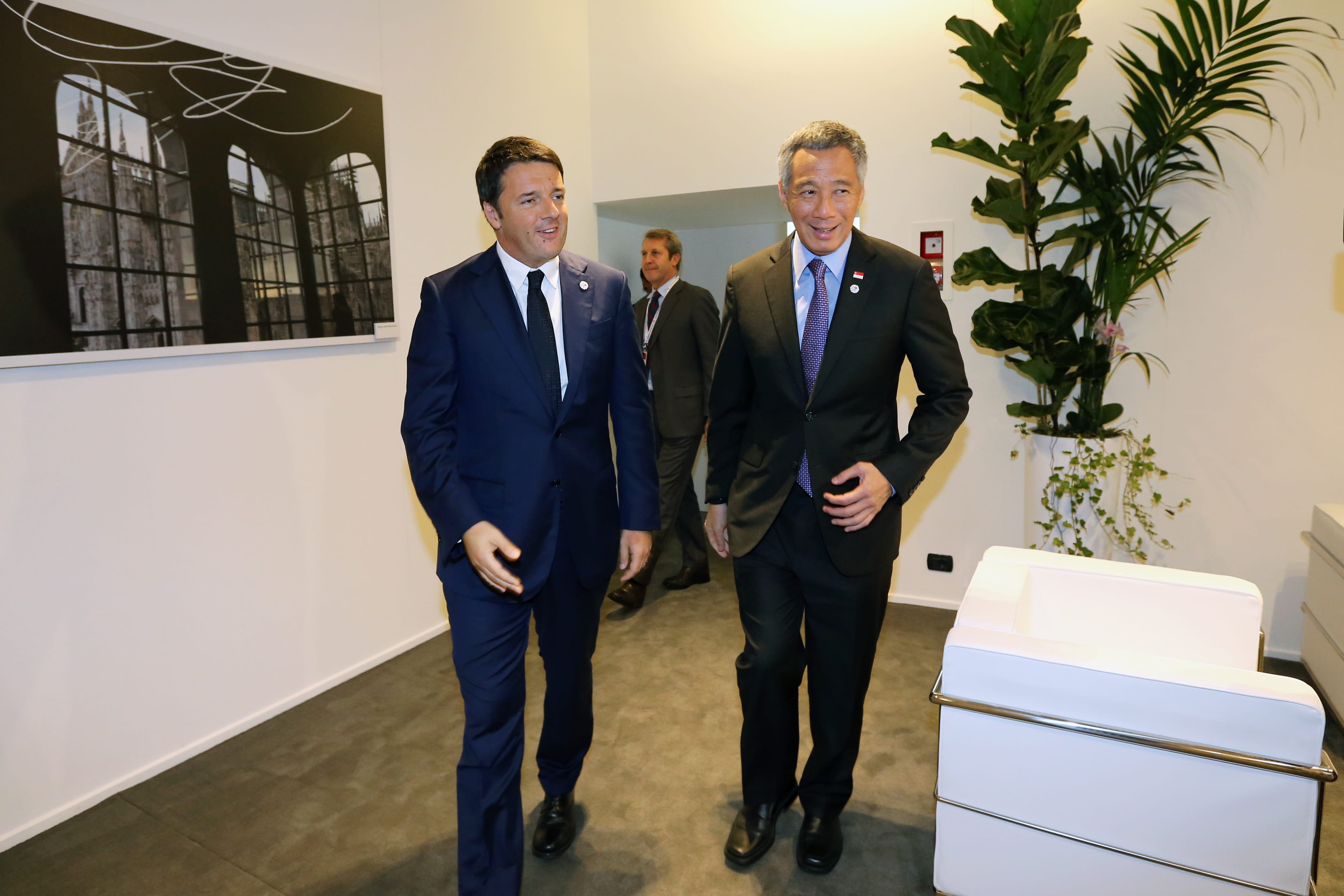 Two men in suits, one Lee Hsien Loong, walk in an interior, near art and plants.